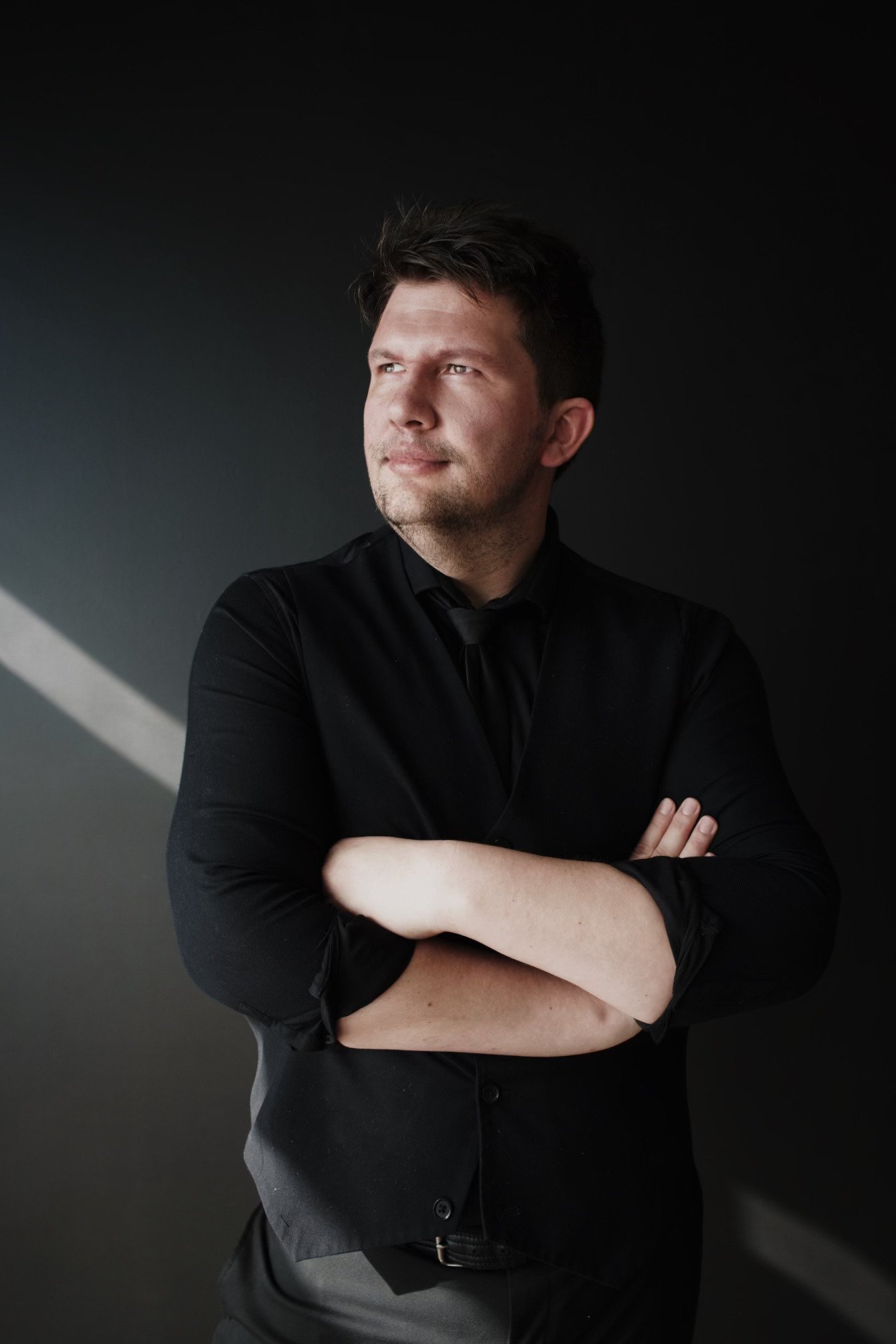 A man with dark hair and light skin wearing a black shirt and black tie stands against a dark background with a diagonal light beam.