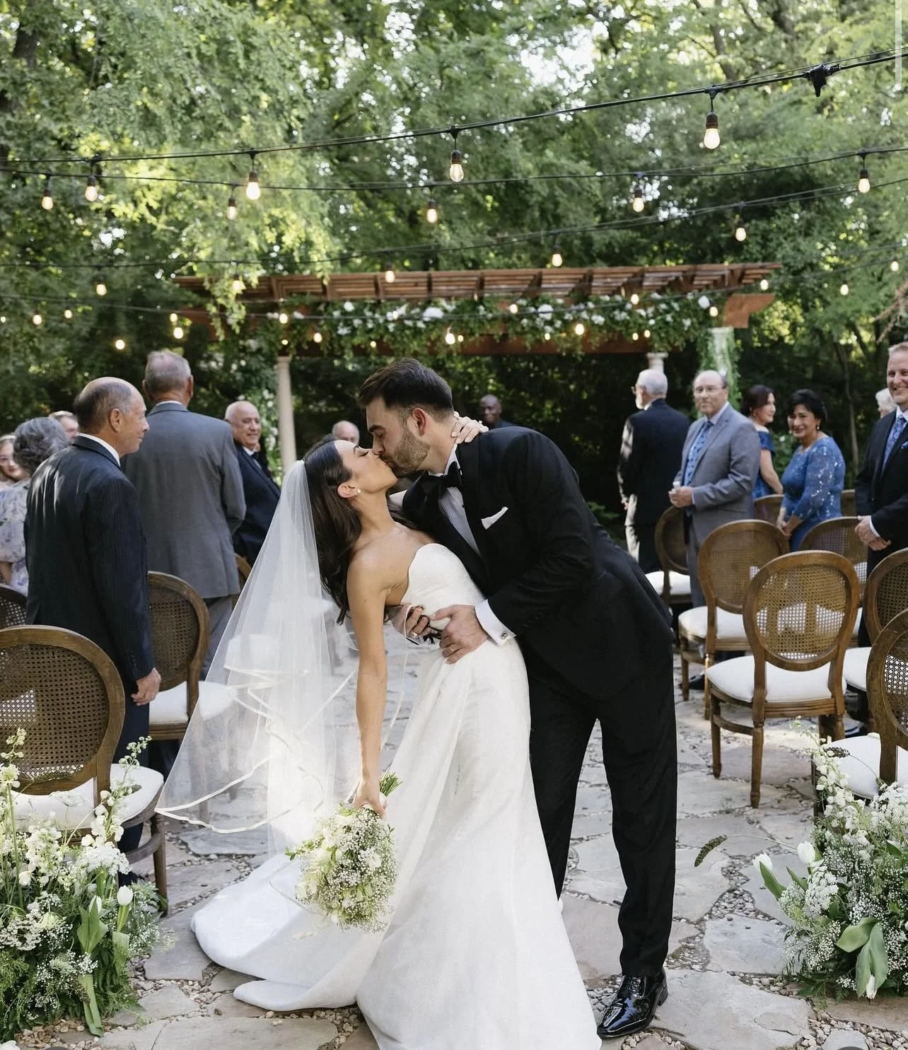 A bride and groom kiss at their outdoor wedding ceremony, surrounded by seated guests and decorated with string lights and floral arrangements.