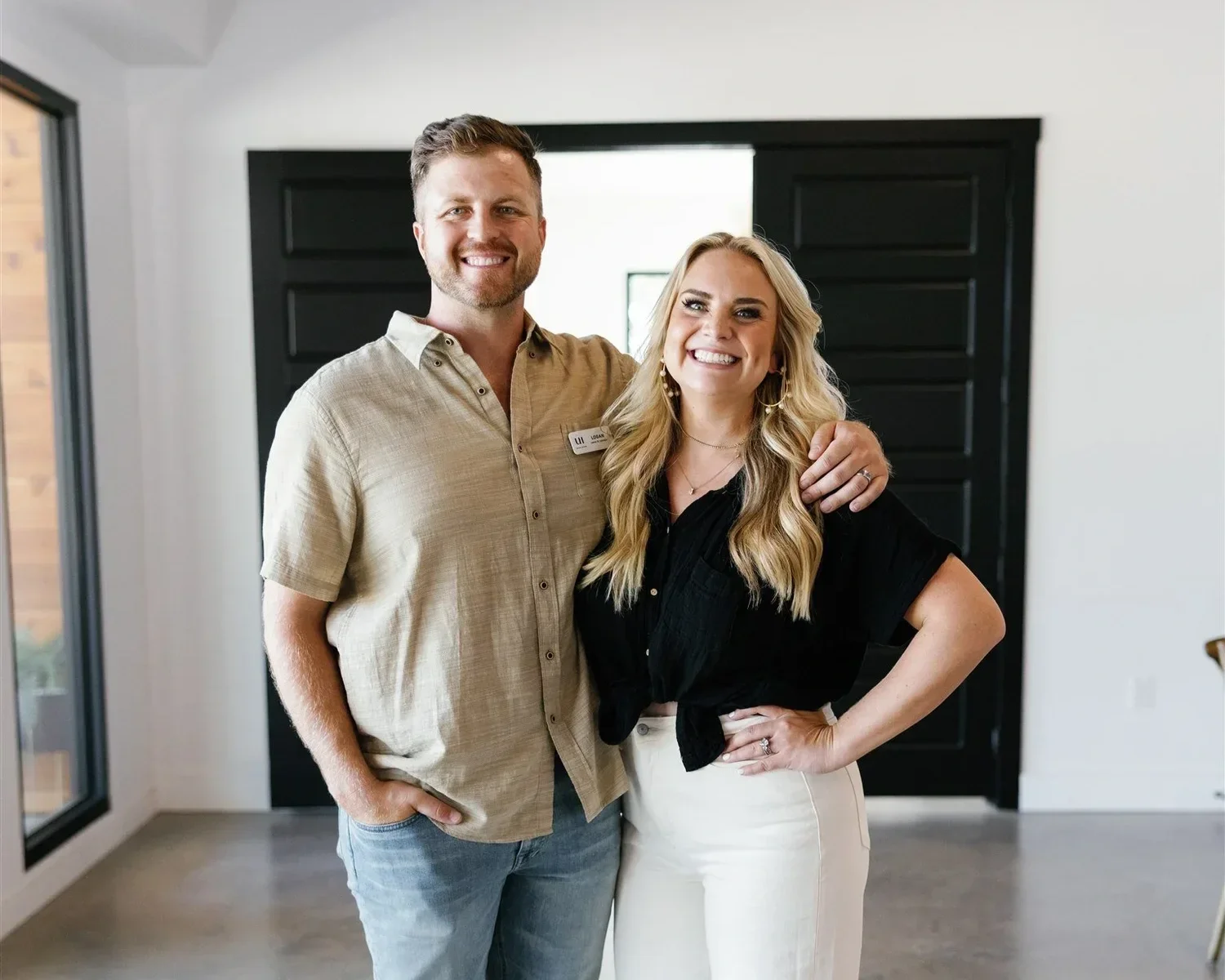 A smiling man and woman standing inside a modern house, with the man's arm around the woman's shoulders. The man has short brown hair, a beard, and is wearing a beige short-sleeve button-up shirt and jeans. The woman has long blonde hair, large hoop earrings, and is wearing a black blouse and white pants.