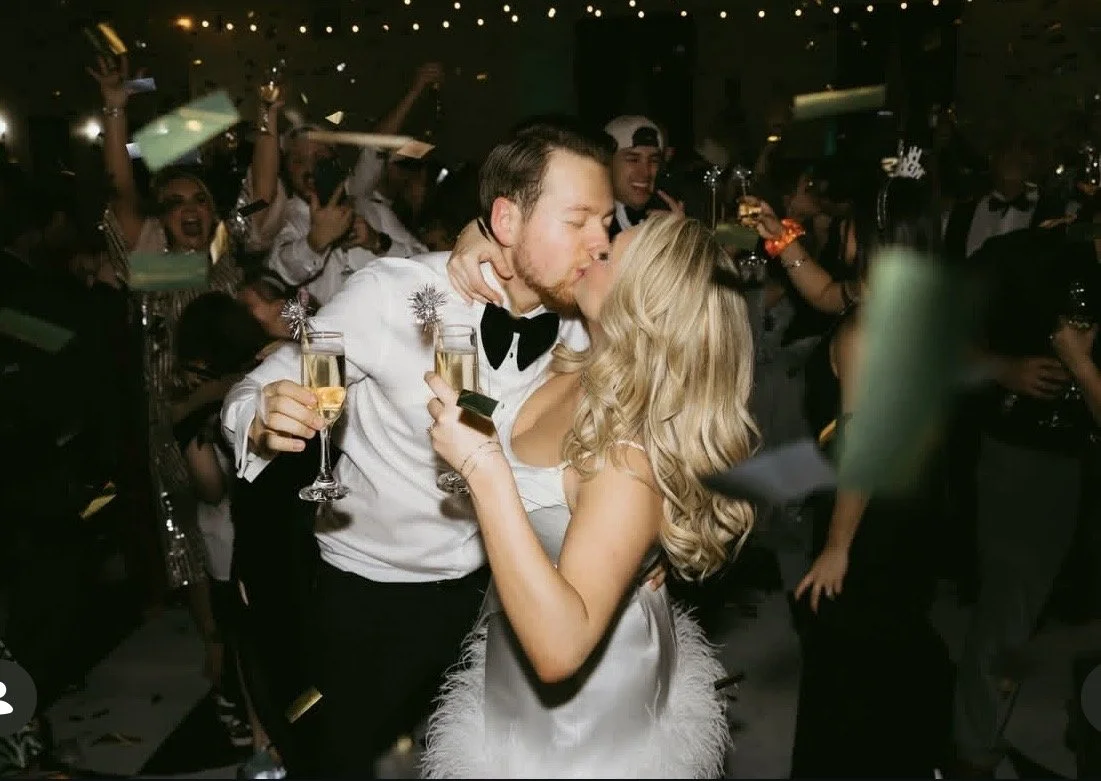 A couple kisses at a celebration with a crowd behind them, holding champagne glasses and toasting, festive lights above.