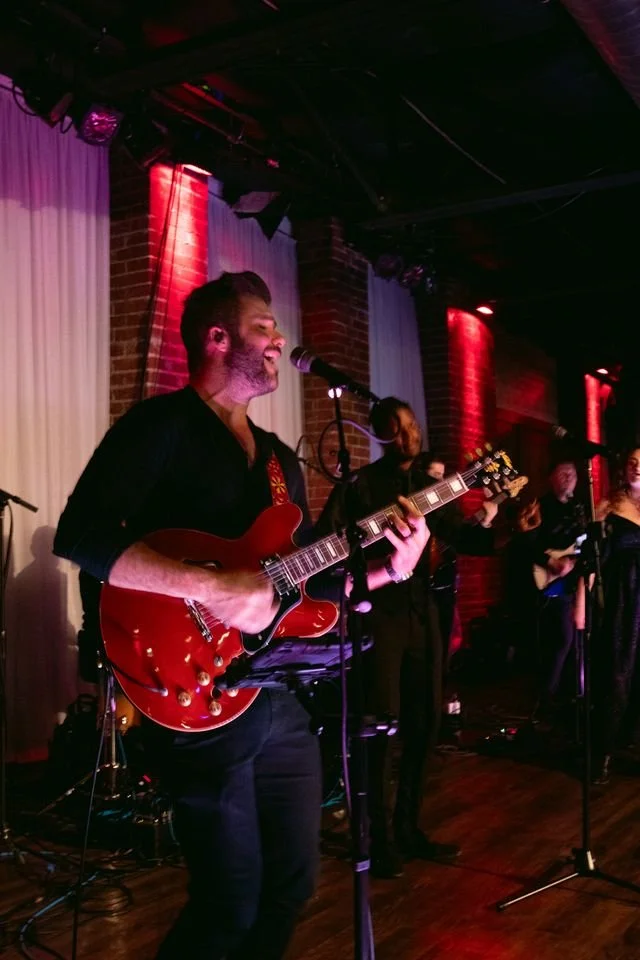 Musicians performing on stage with guitars and microphones in a dimly lit venue with brick walls and red lighting.