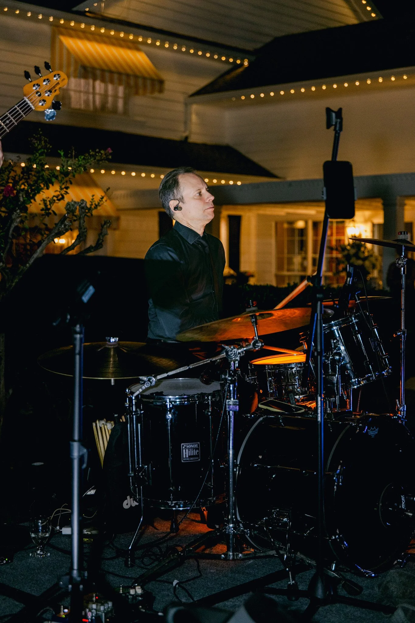 A man playing drums at night during an outdoor event, with house lights and string lights overhead.