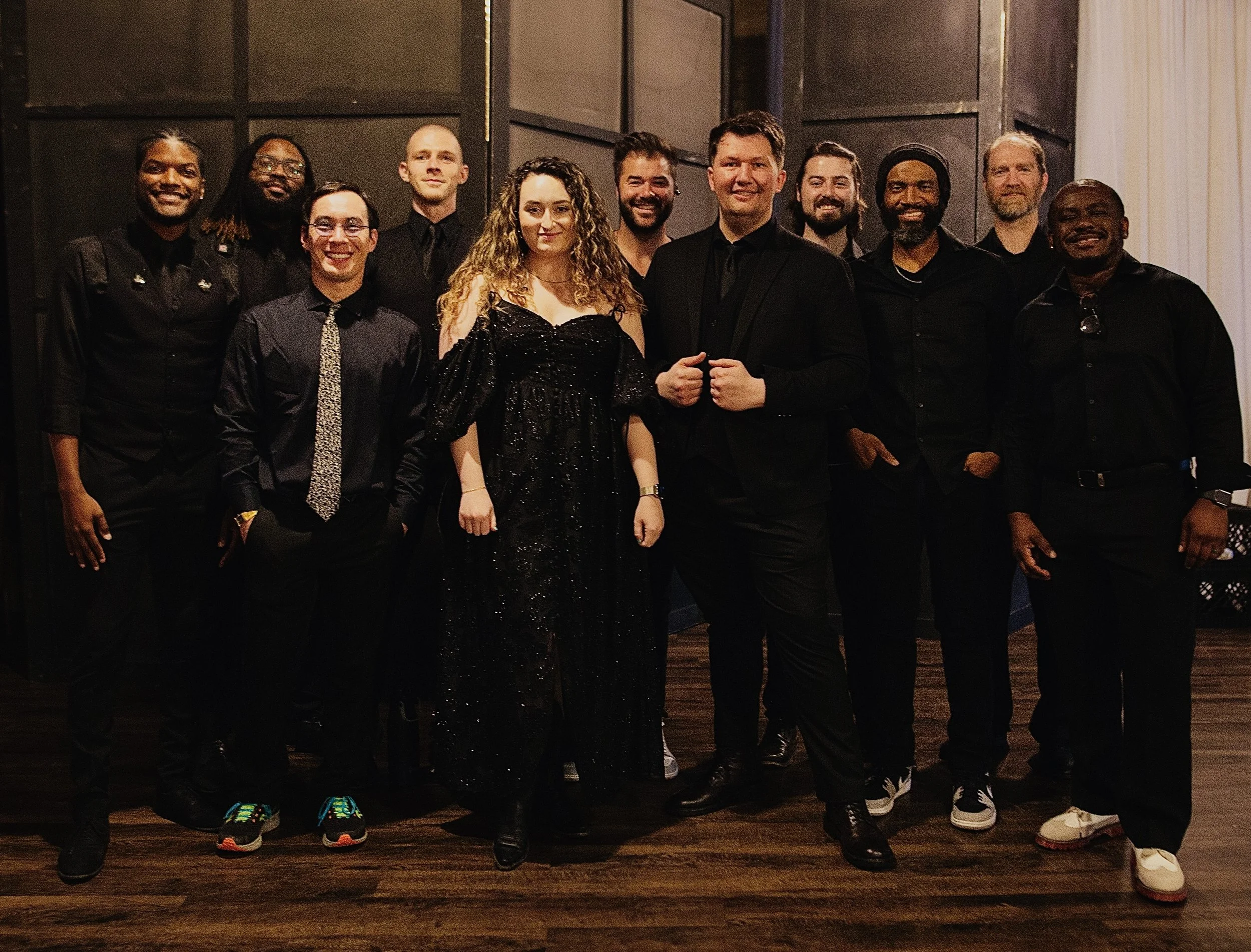 A group of thirteen people dressed in formal black attire, posing indoors against a dark background, smiling for a group photo.
