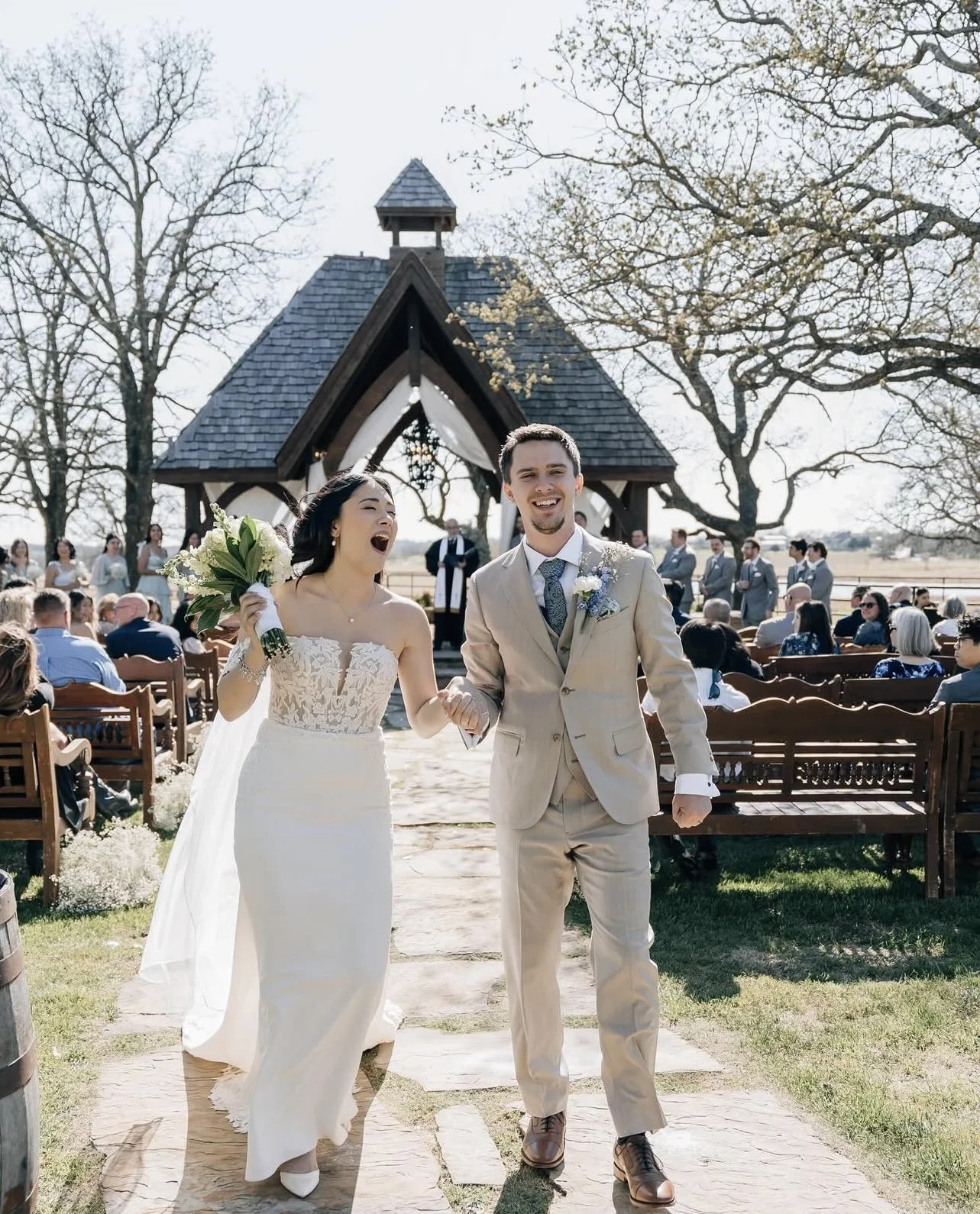 A newlywed couple walking down the aisle after their wedding ceremony outdoors, with the bride holding a bouquet and both smiling, surrounded by seated guests and a small chapel in the background under a clear sky.