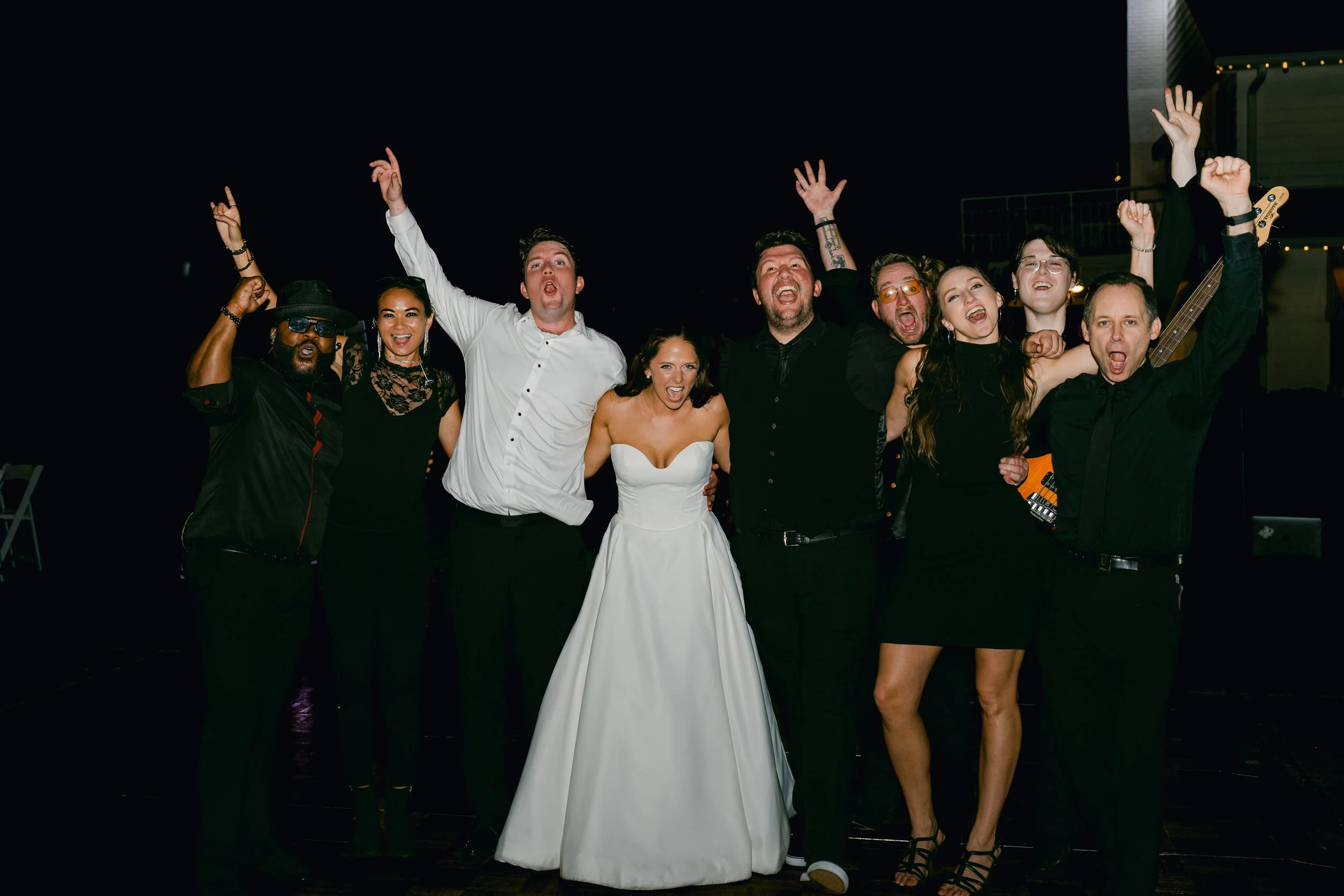 Group of people celebrating at a wedding reception, posing for a photo with arms raised and smiling. The bride is in a white wedding dress, and others are dressed in black and white attire, with some holding musical instruments.