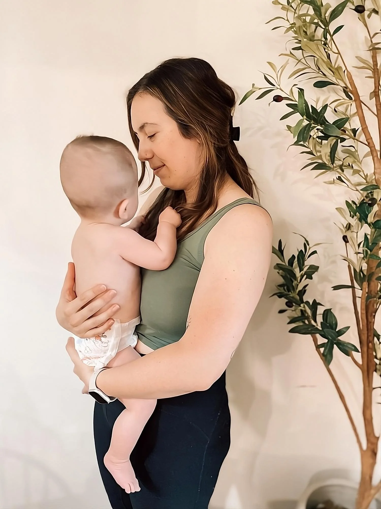 A woman holding a young child with no shirt and a diaper, both with eyes closed and forehead touching, standing indoors near a potted plant.