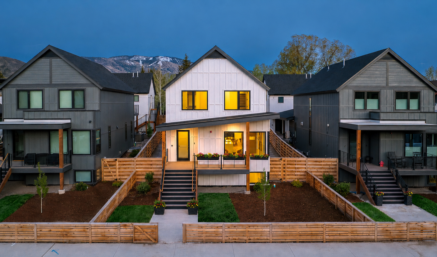 A row of modern townhouses with front porches, some lit from within, set against a mountain backdrop at dusk, with landscaped front yards.