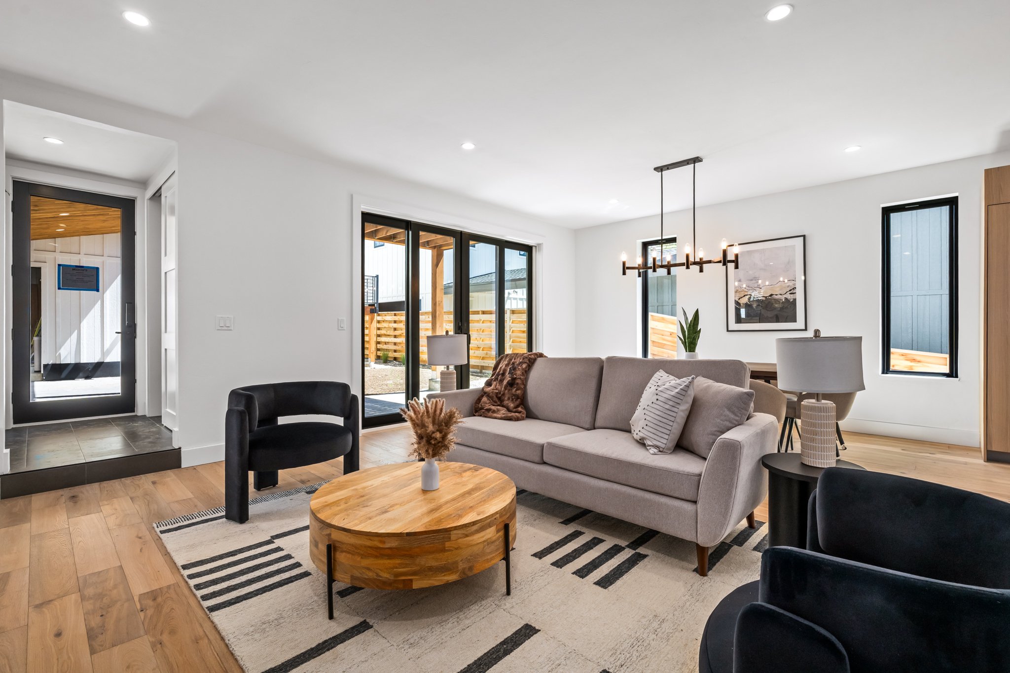 Open-concept living room with beige sofa, black accent chairs, wooden coffee table, large sliding glass doors, modern chandelier, and framed artwork, with wood flooring and white walls.