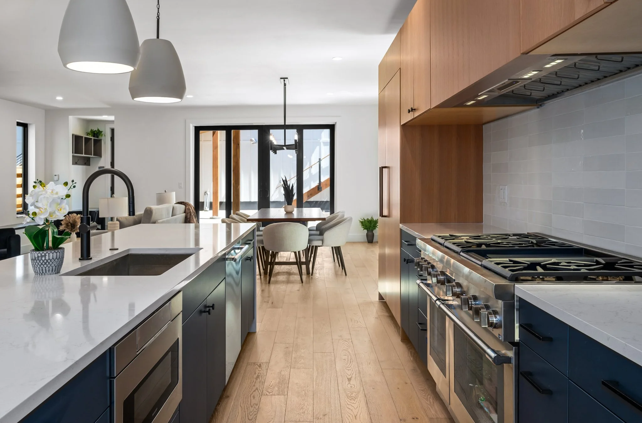 Modern kitchen with island counter, black lower cabinets, stainless steel stove, wooden upper cabinets, beige tile backsplash, and dining area with table and chairs visible in the background at Eighth Street Homes Steamboat