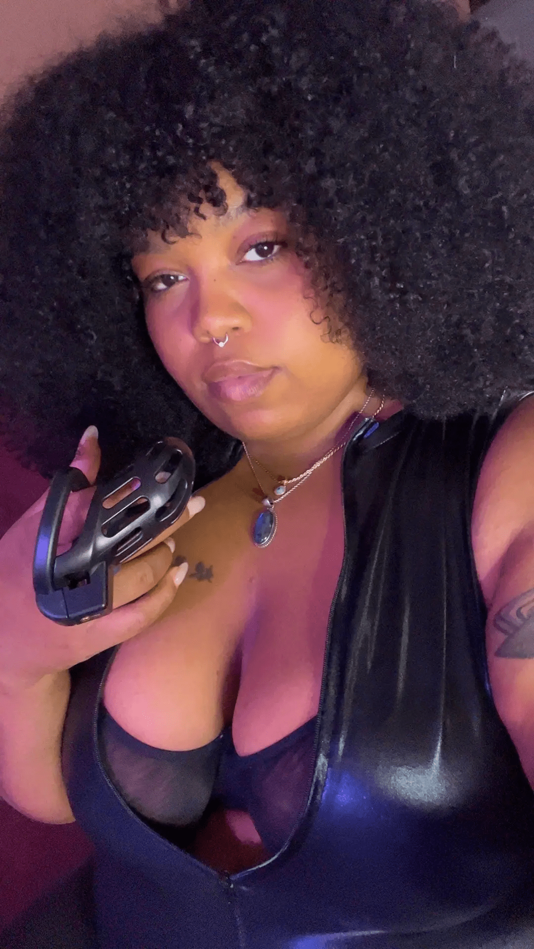 Mistress Khai with curly black hair, wearing a black leather top and jewelry, holding a black chastity cage.