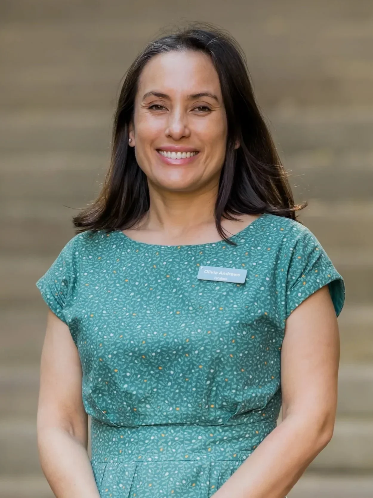 A woman with long dark hair smiling, wearing a teal patterned dress and a name tag that reads Olivia Andrews, in front of a blurred neutral background.
