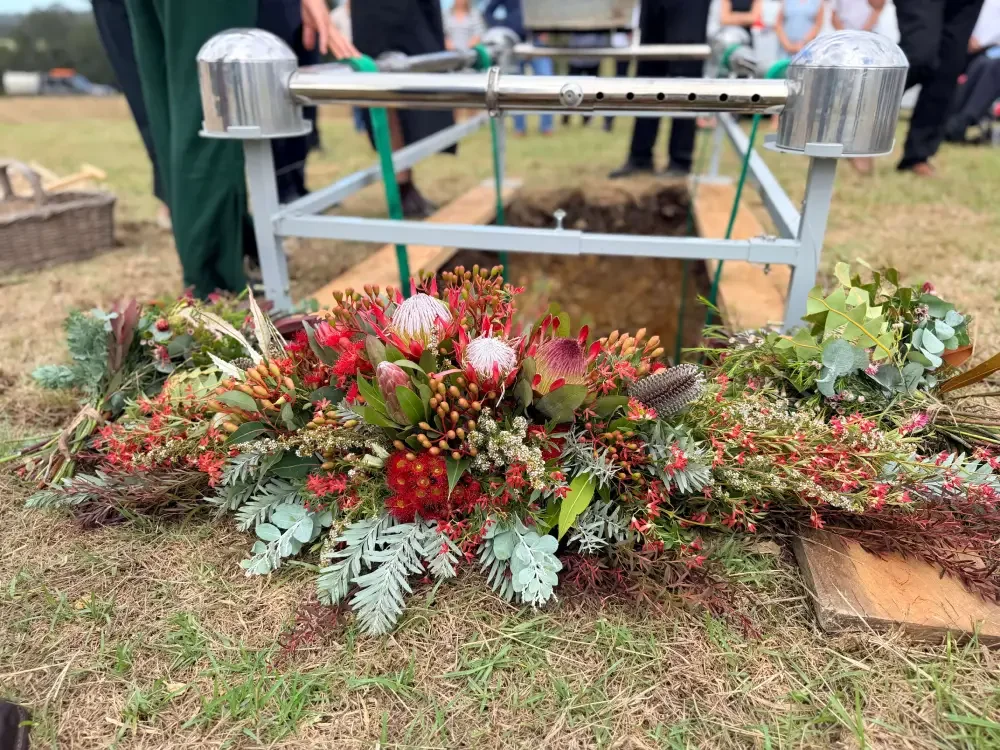 A floral arrangement laid around a grave site with a coffin underneath, during a funeral service outdoors.