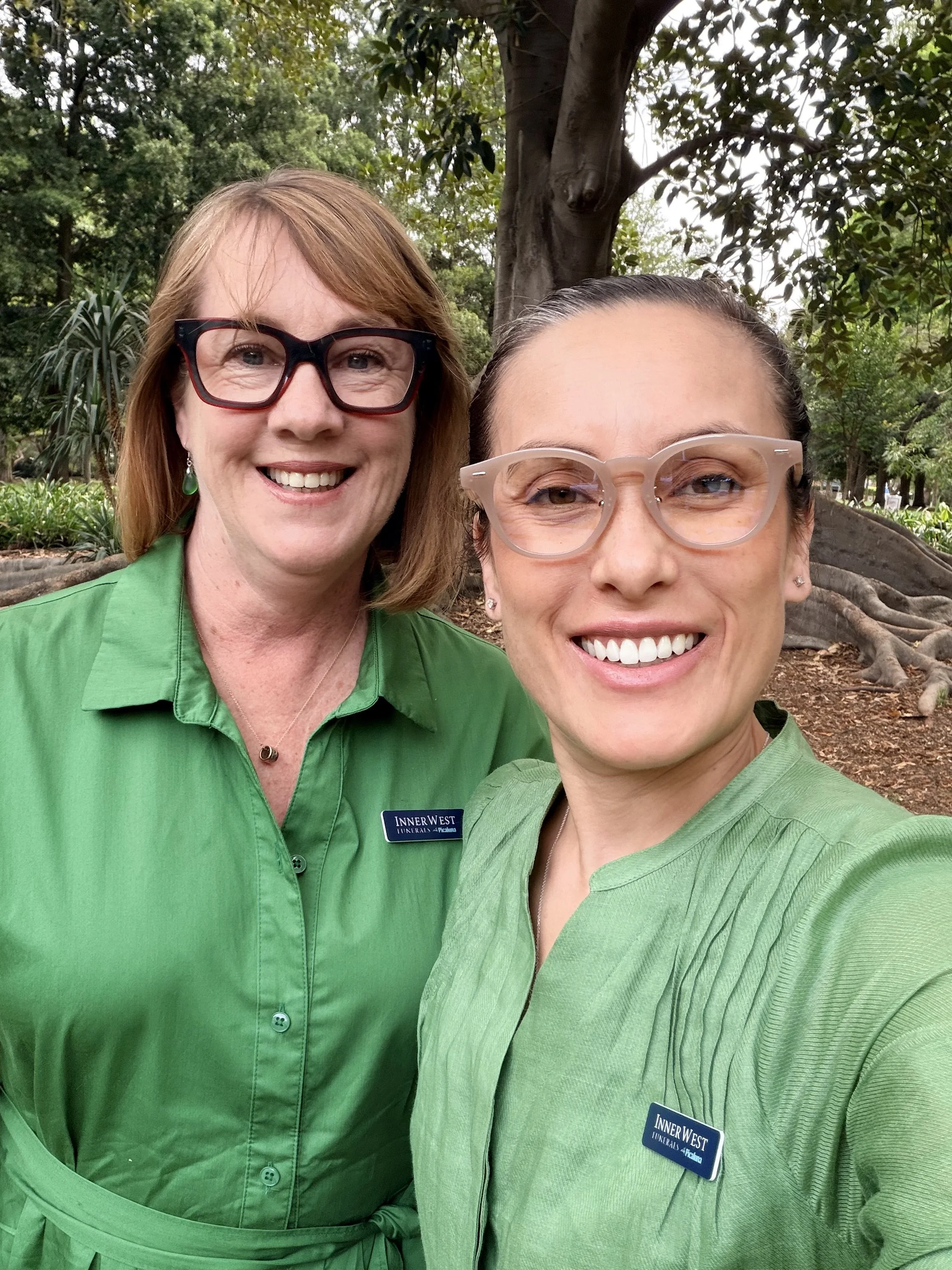 Two women taking a selfie outdoors, both wearing green shirts and glasses, with a large tree and green foliage in the background.