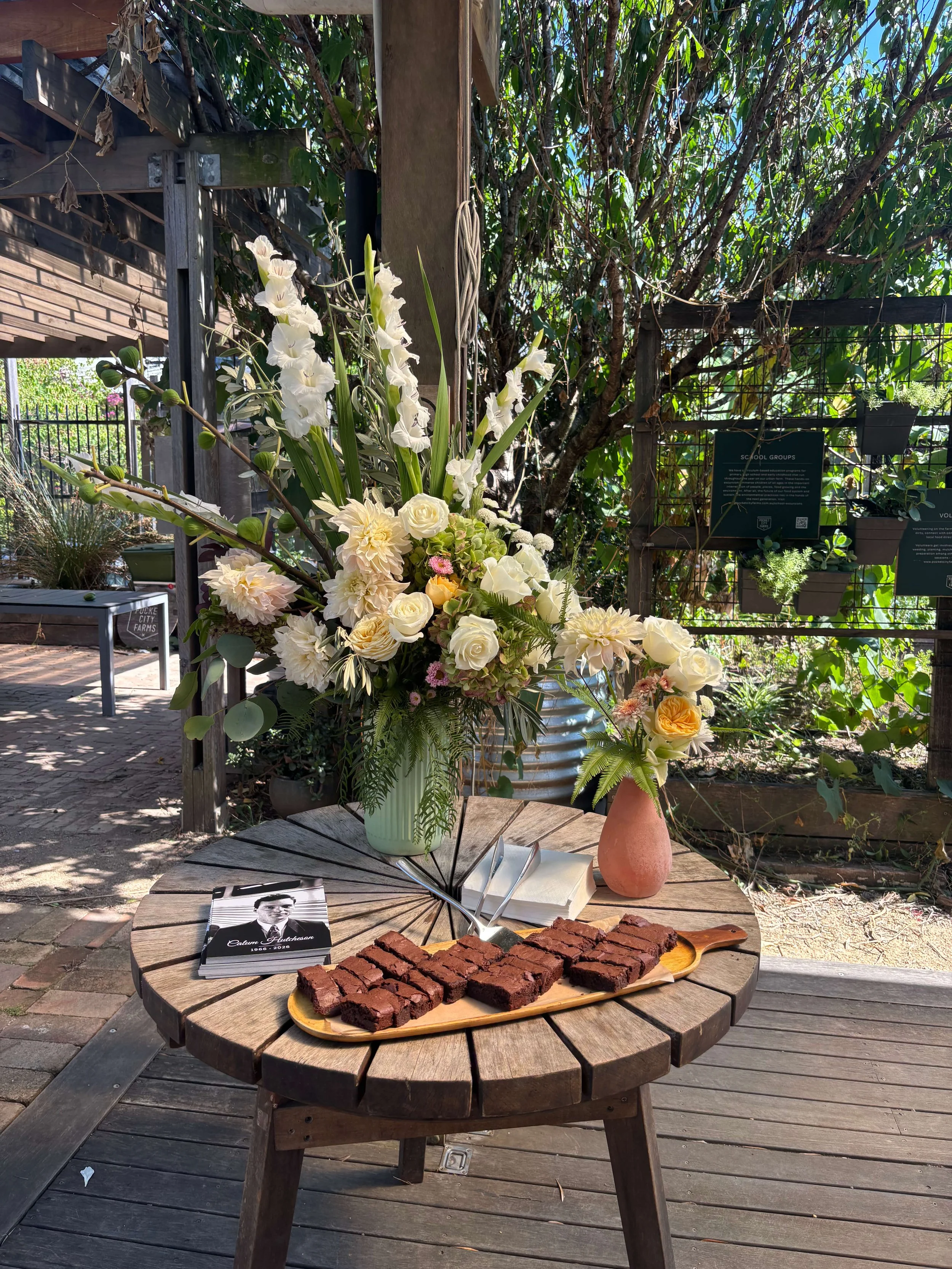 A round wooden table outdoors with a vase of white flowers, a smaller peach-colored vase, a black-and-white booklet, and a plate of brownies. In the background, there is greenery, a wooden fence, and a grid rack with potted plants and informational signs.