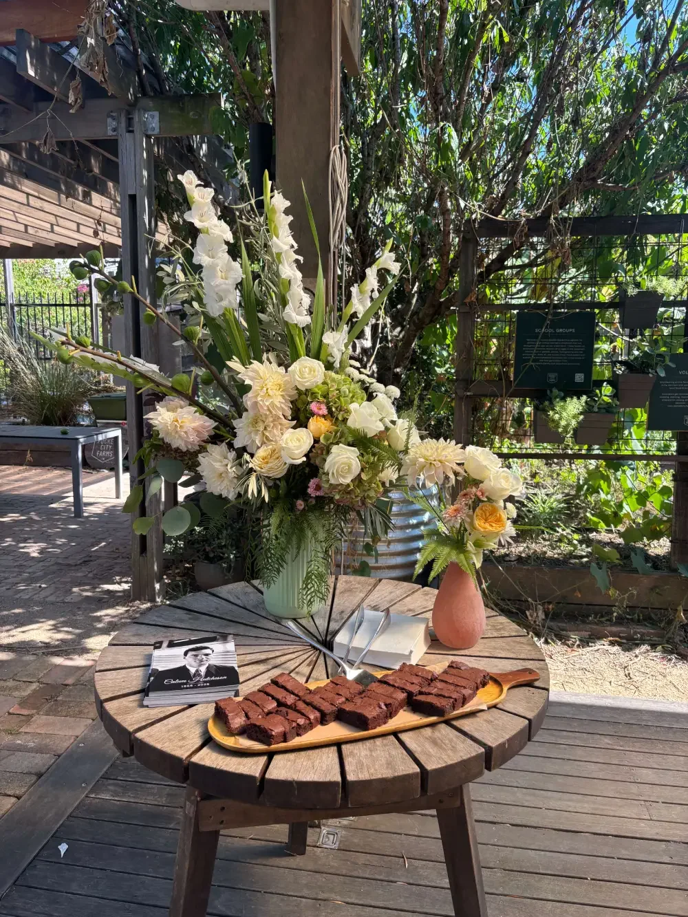A wooden round table with a yellow tray of brownies, a book, and a pair of tongs, two vases with white and yellow flowers, and a small stack of napkins, set outdoors with plants and trees in the background.