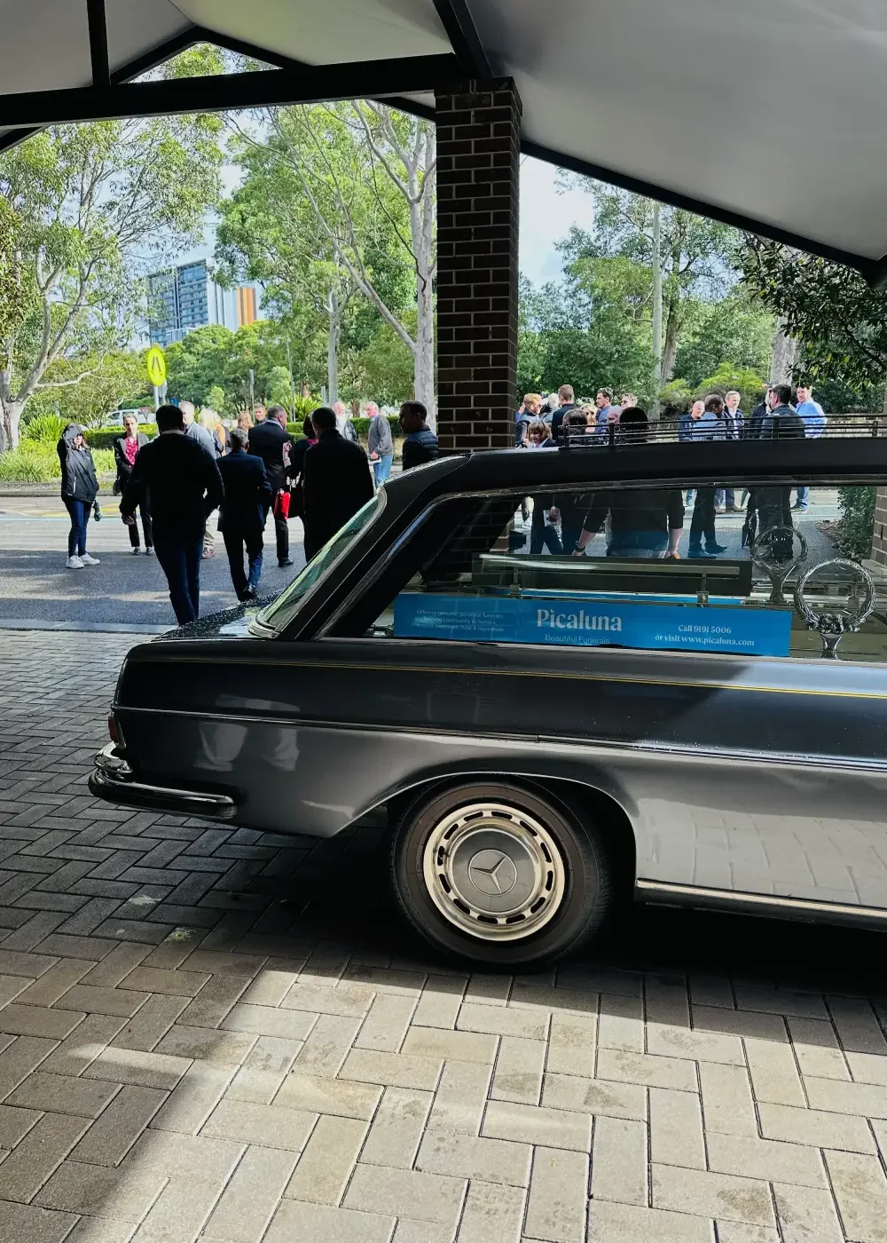 A vintage black Mercedes-Benz parked outside a building with a group of people gathering at a bus stop or outdoor seating area, surrounded by trees and urban buildings in the background.
