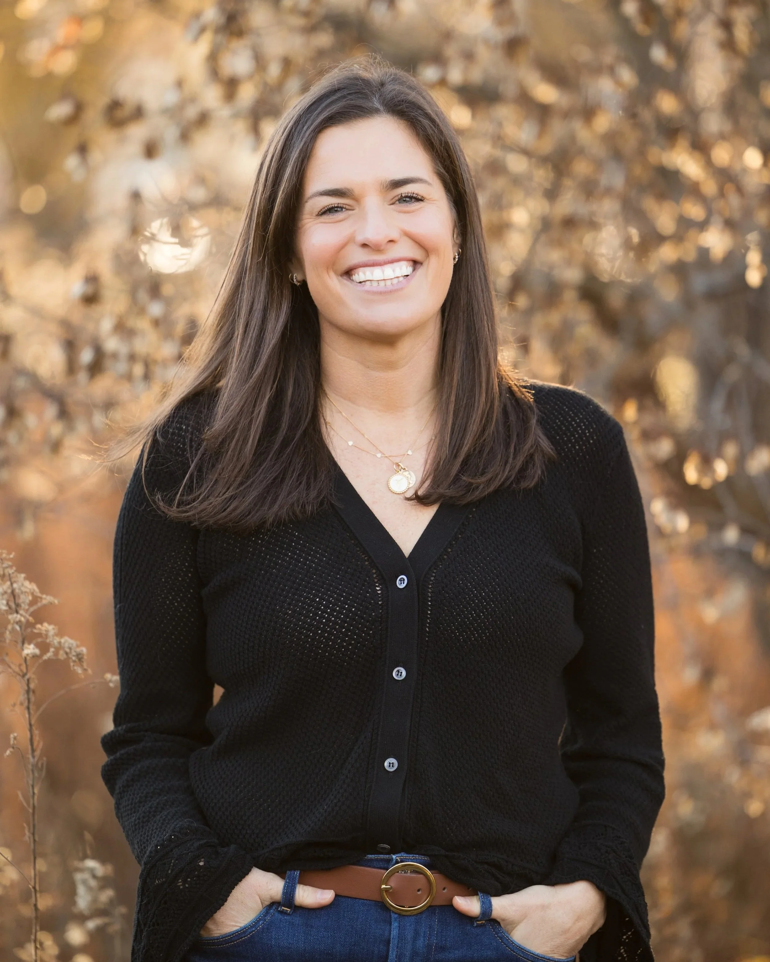A woman smiling outdoors during autumn, wearing a black sweater, blue jeans, and a gold necklace, with her hands in her pockets.
