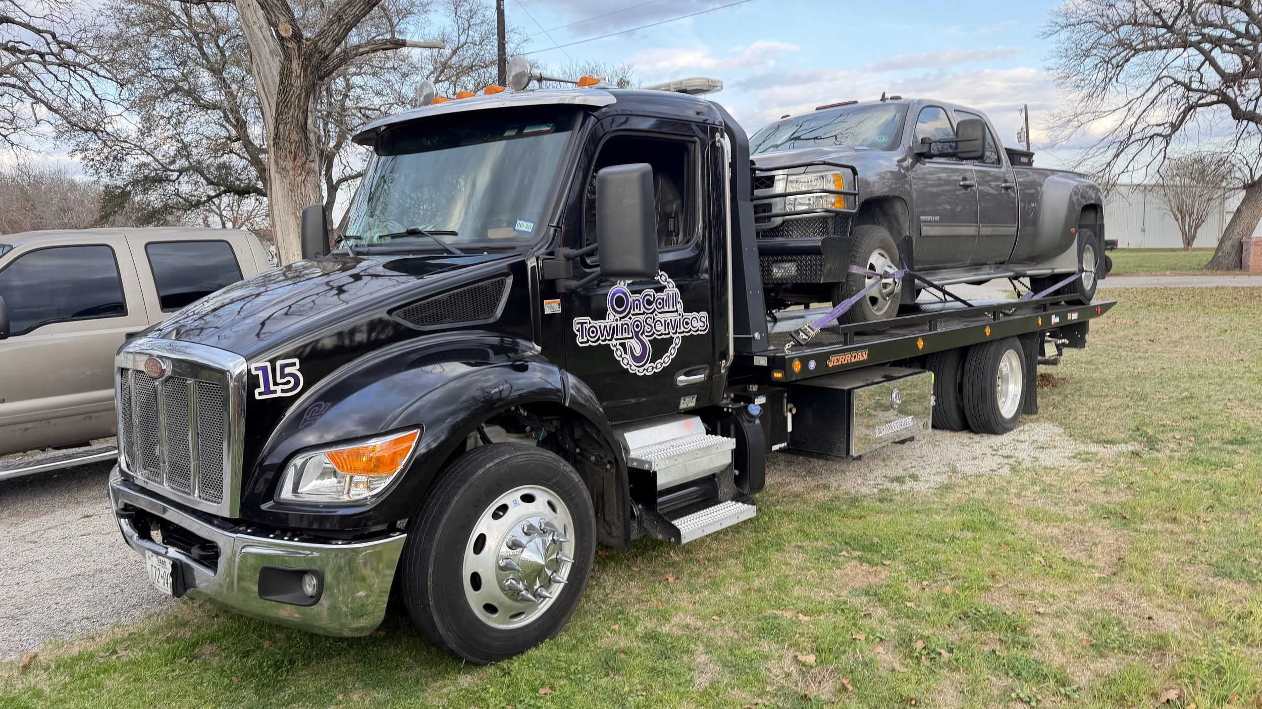 A black tow truck carrying a dark gray pickup truck on its flatbed, parked on grass with trees and a building in the background.