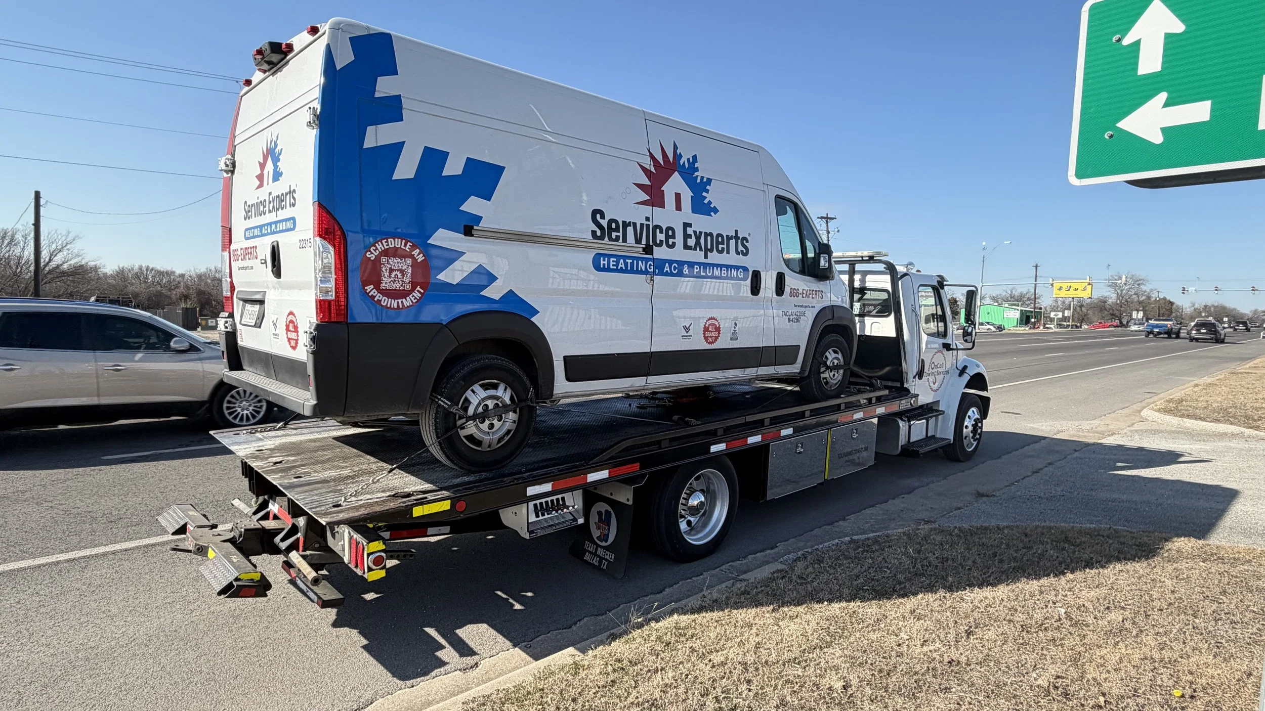 A Service Experts heating, AC, and plumbing vehicle being transported on a flatbed truck on the side of a busy road.