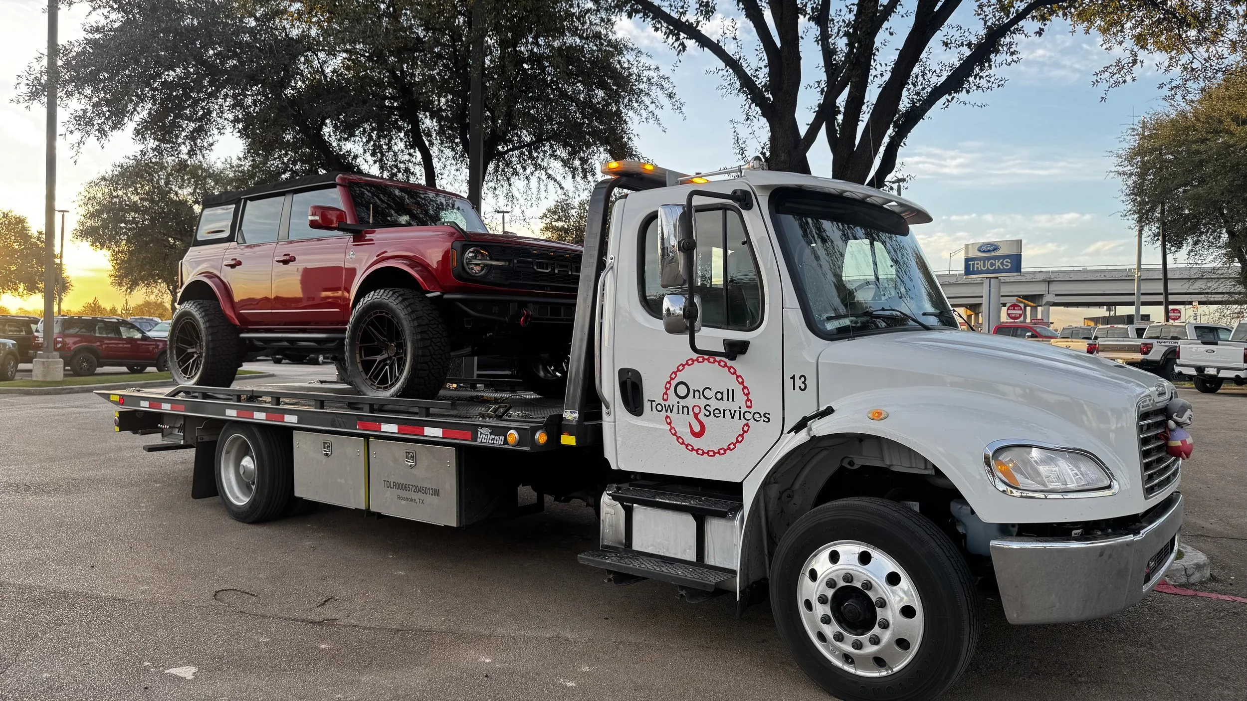 A white tow truck carrying a red and black SUV in a parking lot during sunset.