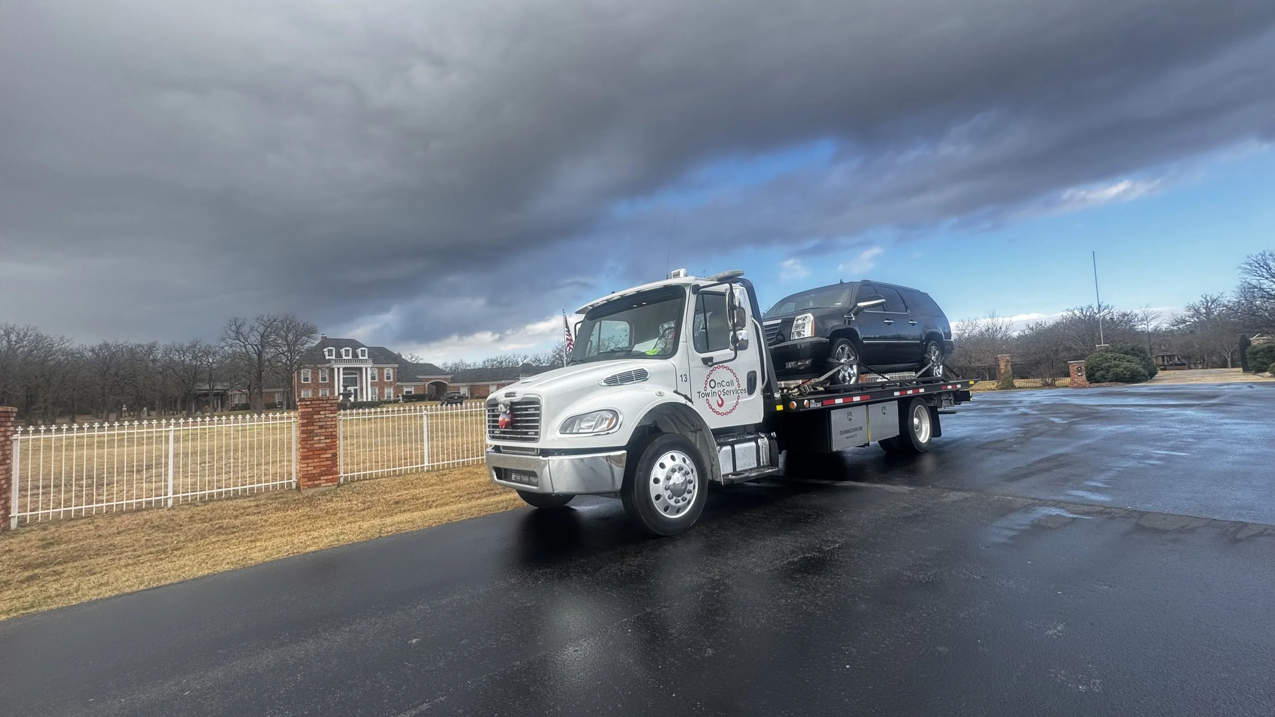A flatbed tow truck carrying a black SUV parked on a wet asphalt surface with dark clouds overhead and a large house with a brick and white fence in the background.