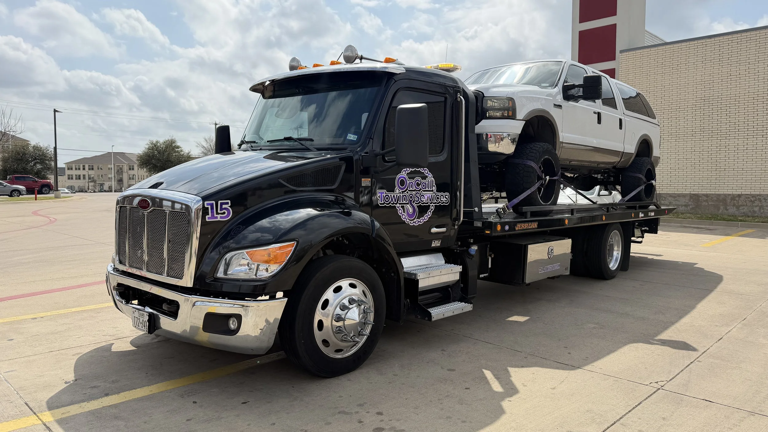 A black tow truck with purple lettering on the door that reads 'OnCall Towing Services' and the number 15. The truck is transporting a white pickup truck on its flatbed.