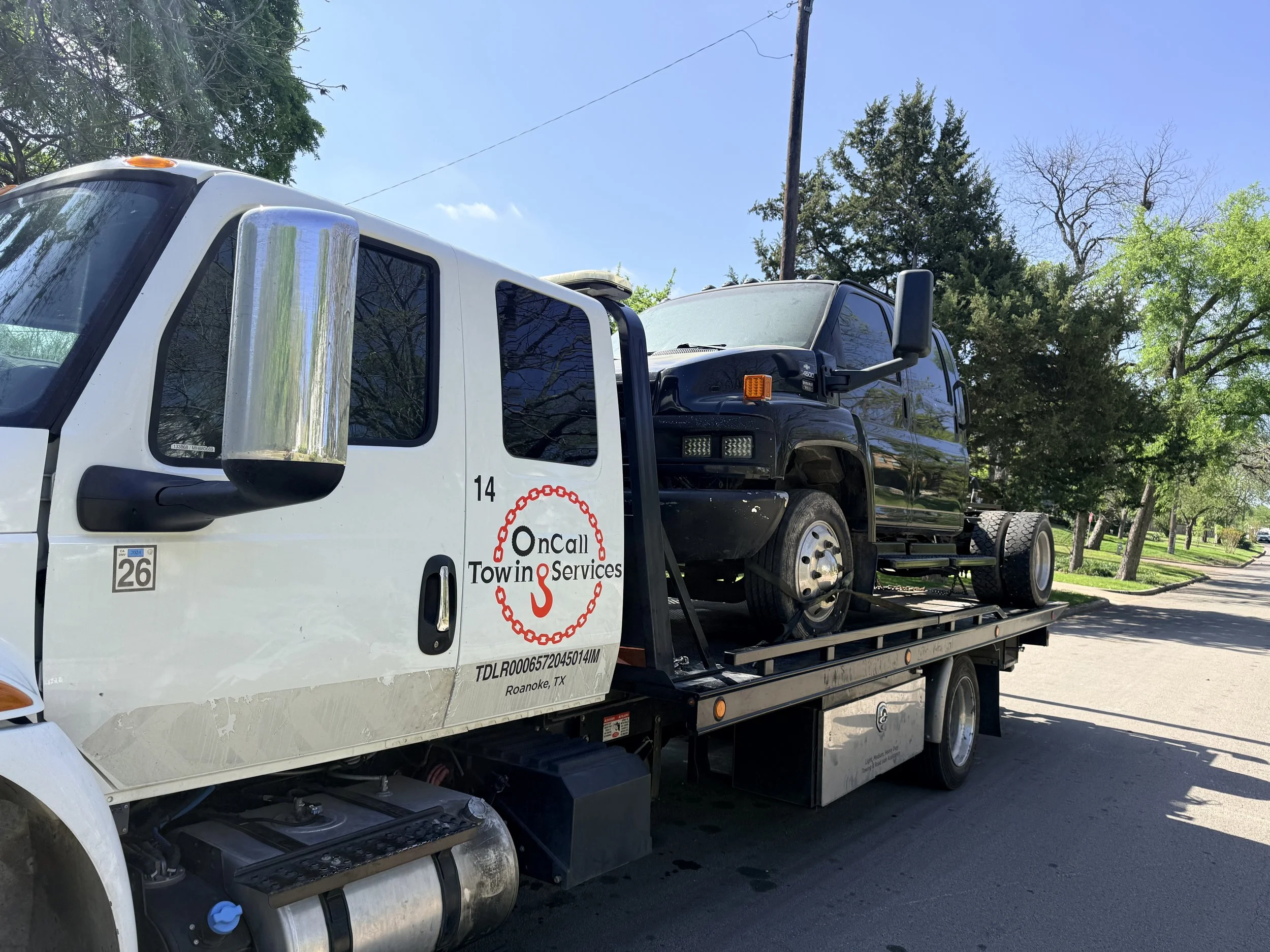 Tow truck carrying a black vehicle on a sunny street with trees and houses.