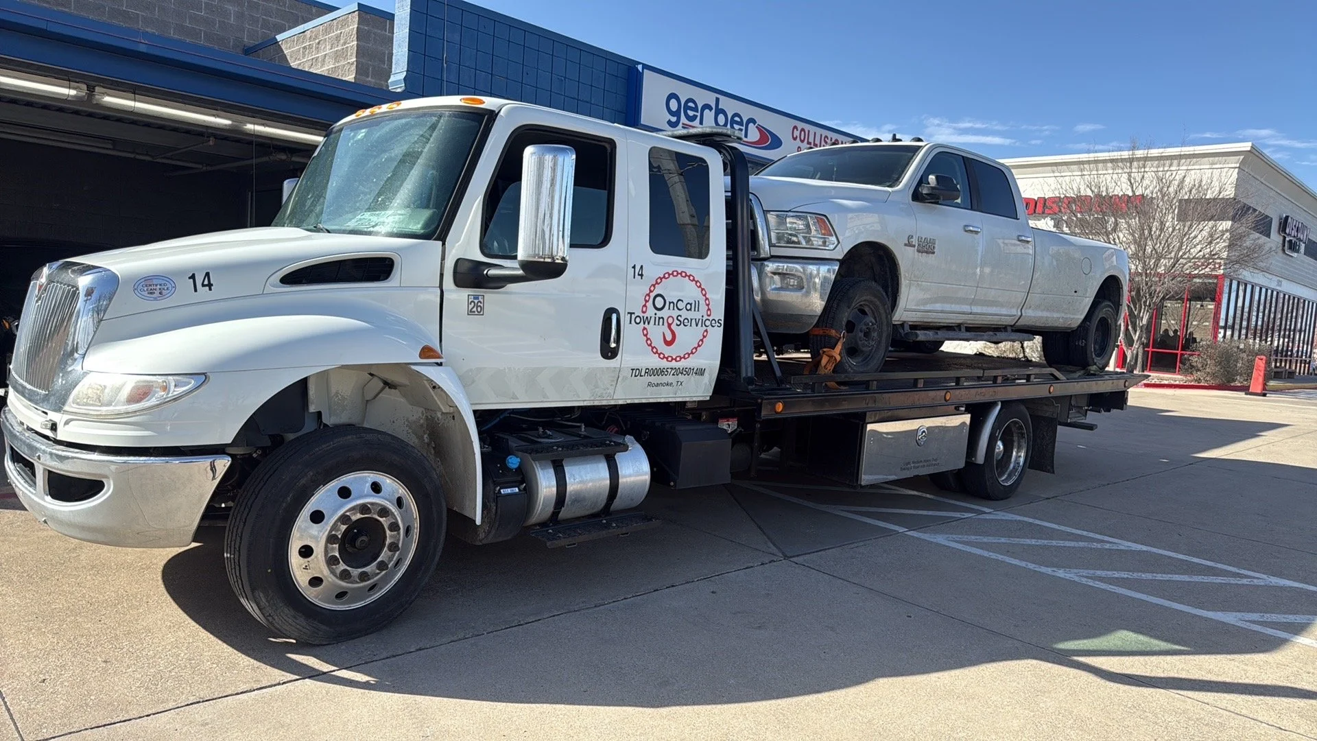Flatbed tow truck carrying a silver pickup truck, parked outside a shopping center on a clear day.