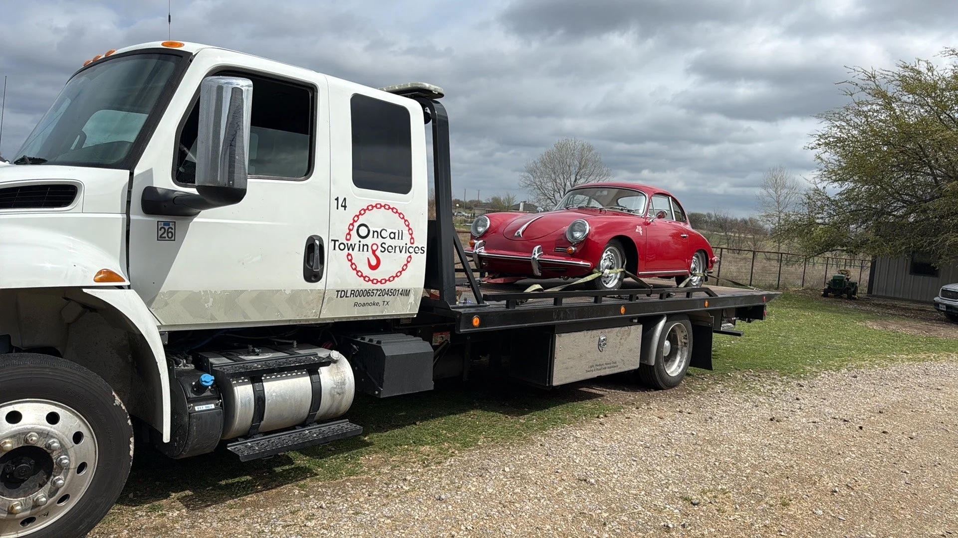 A white tow truck carrying a vintage red car on its flatbed, outdoors on a cloudy day with trees and a gravel ground in the background.
