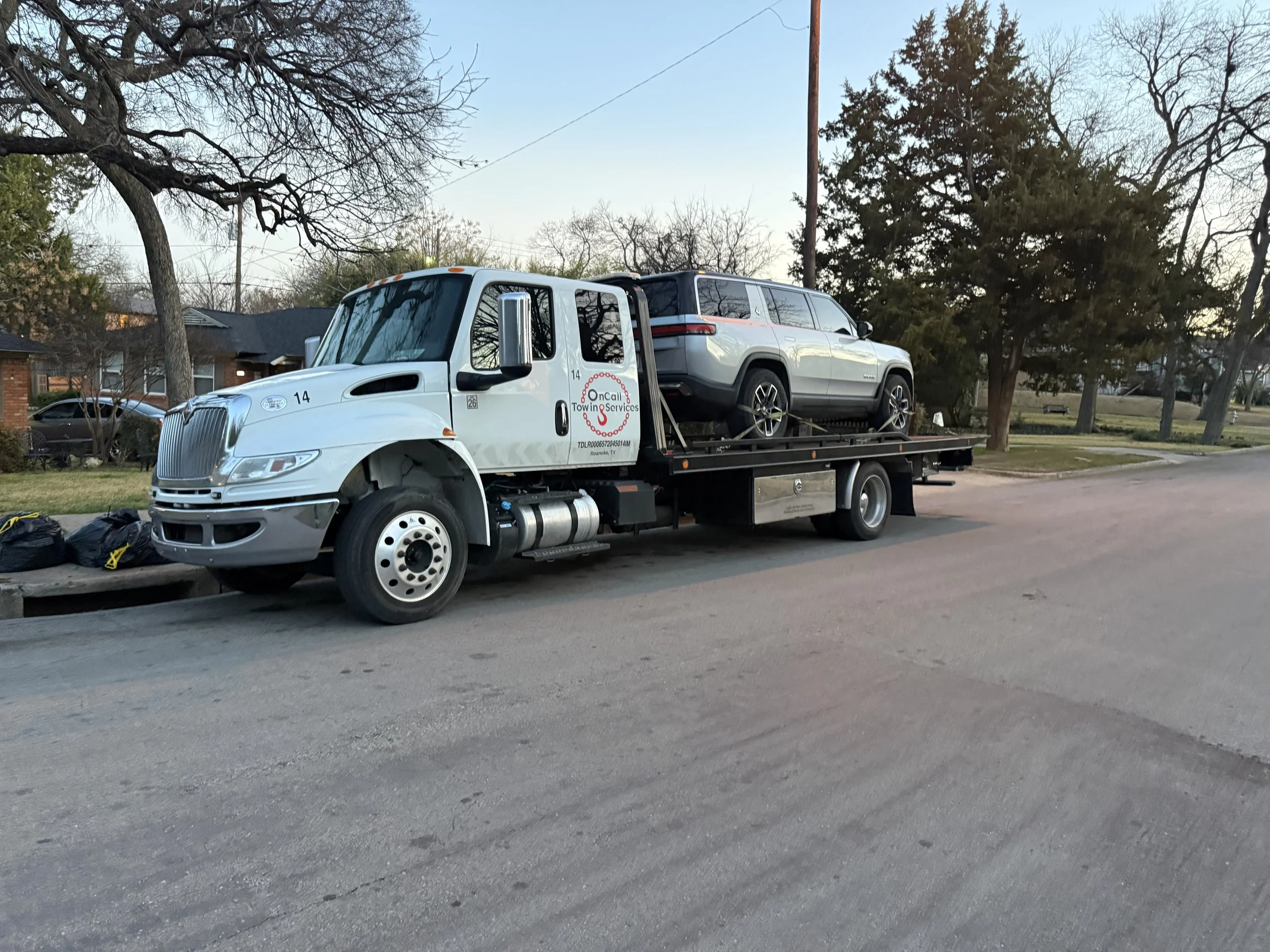Tow truck parked on the side of the street with a silver SUV loaded on its flatbed, in a residential neighborhood with trees and houses in the background during early evening.
