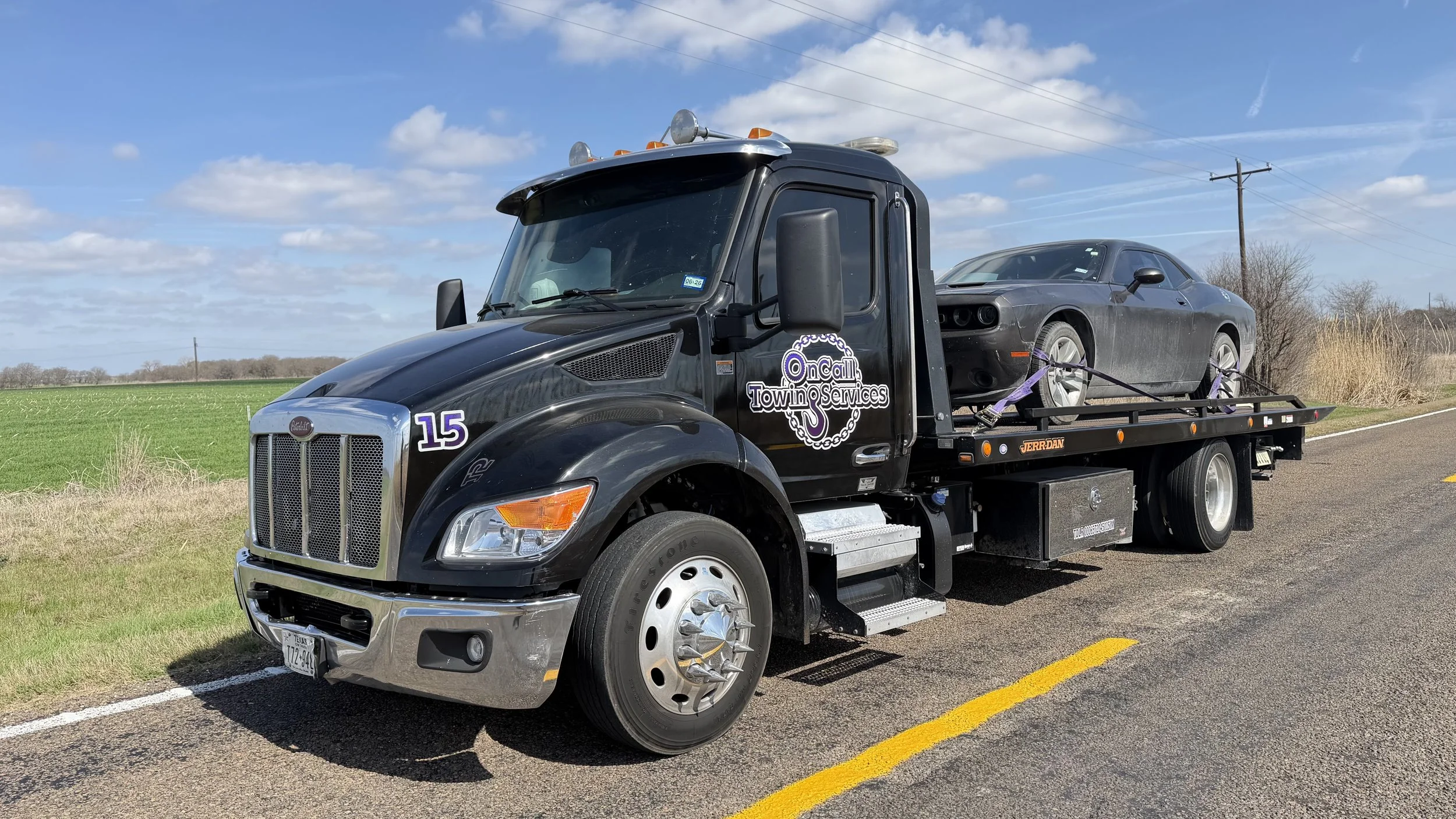 Black tow truck with a silver Porsche car loaded on the flatbed, parked on the side of a rural road under a partly cloudy sky.