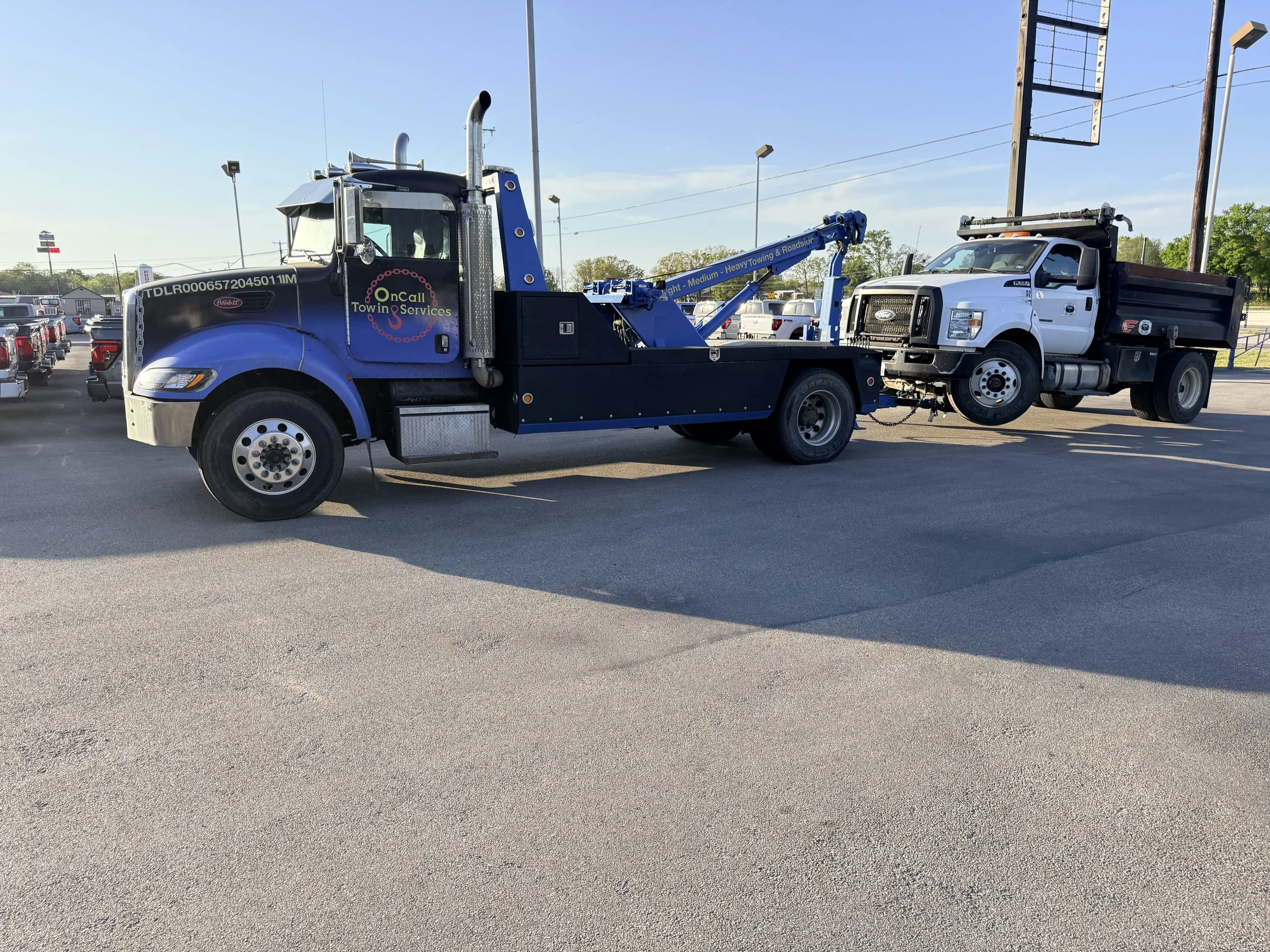 A blue tow truck, labeled 'On Call Towing & Services,' is towing a white flatbed pickup truck in a parking lot during daylight. The tow truck has a crane arm extended.