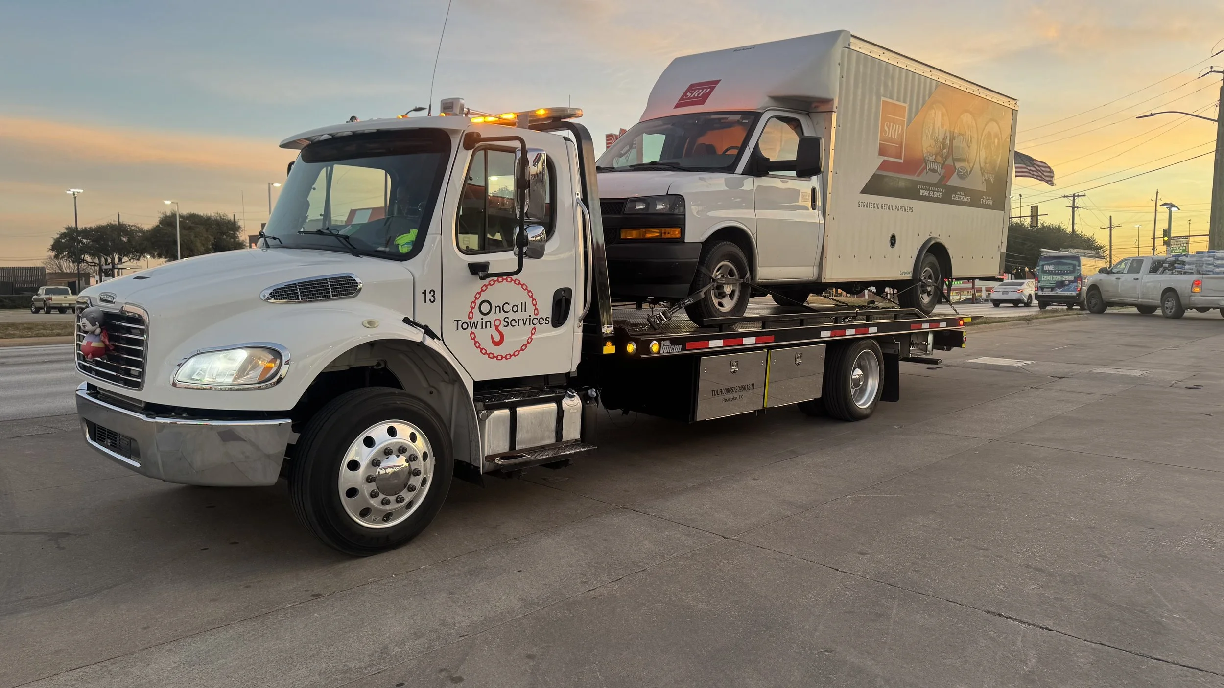 A tow truck carrying a white delivery van with a colorful logo on the side. The scene is set during sunset at a parking lot with other vehicles and streetlights in the background.