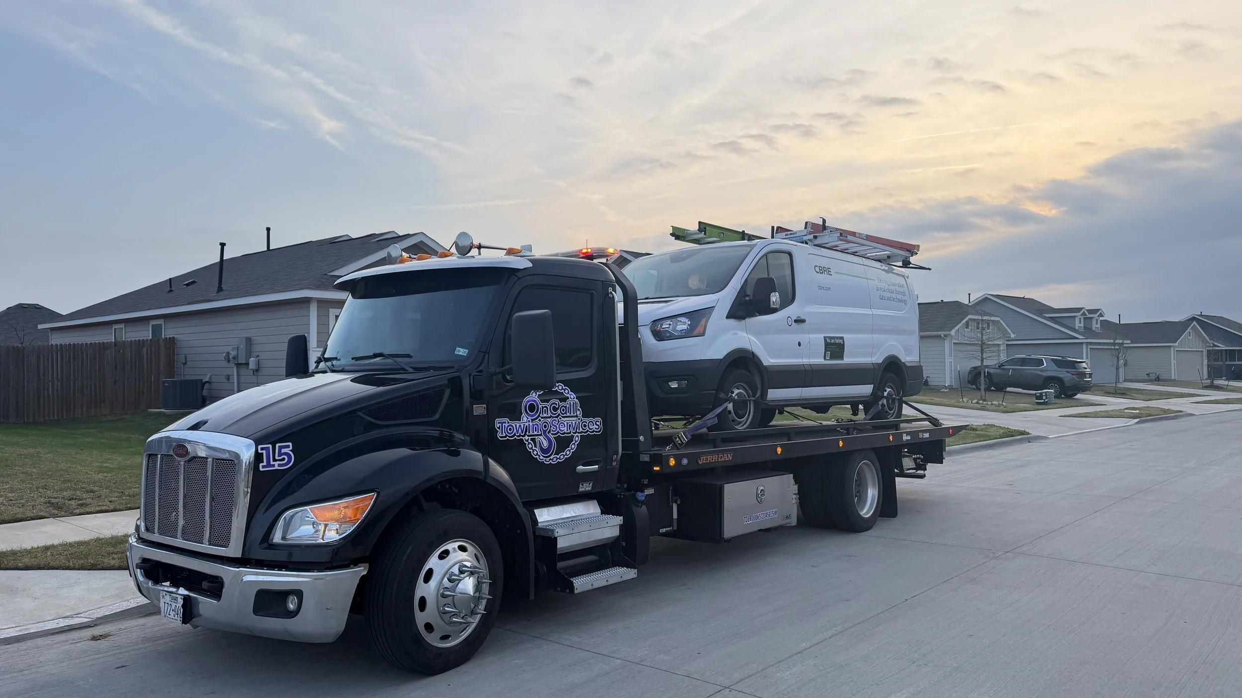 A tow truck is transporting a white utility van with ladders on top, driving through a suburban neighborhood with single-family homes and a few parked cars at dusk.