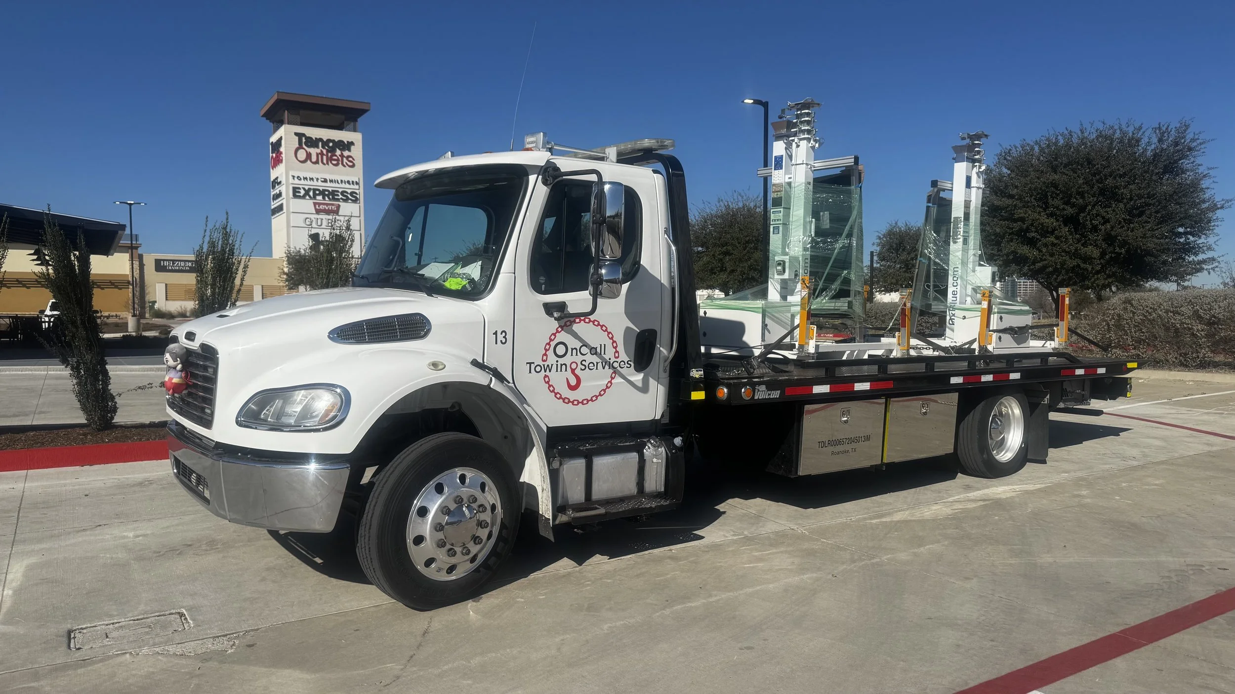 A white tow truck with the logo 'OnCall Tow & Services' parked in a lot, with glass towers on the back and a small plush toy attached to the front grille.