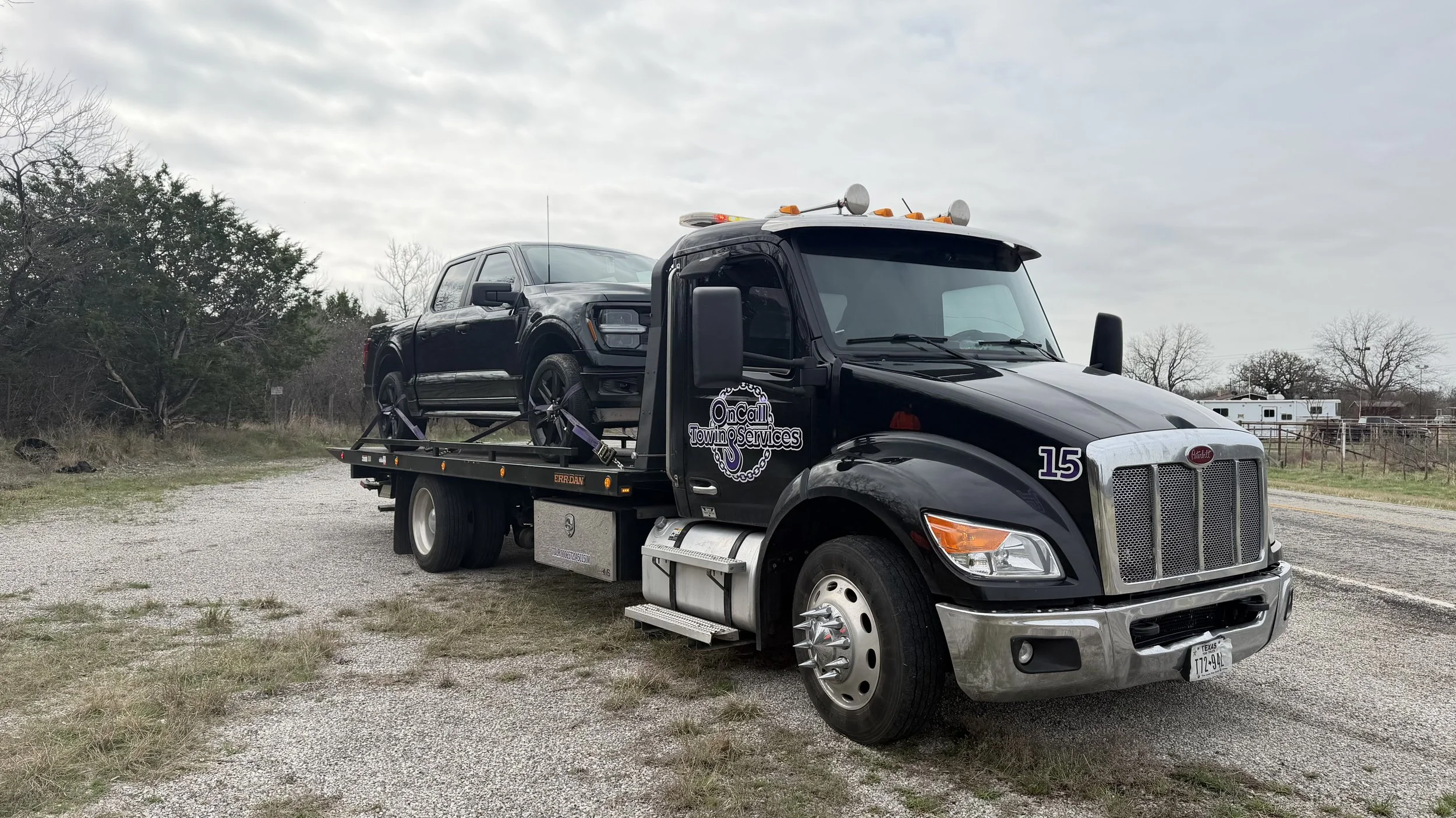 A tow truck from OnCall Towing Services is parked on a gravel lot with a black pickup truck secured on its flatbed.