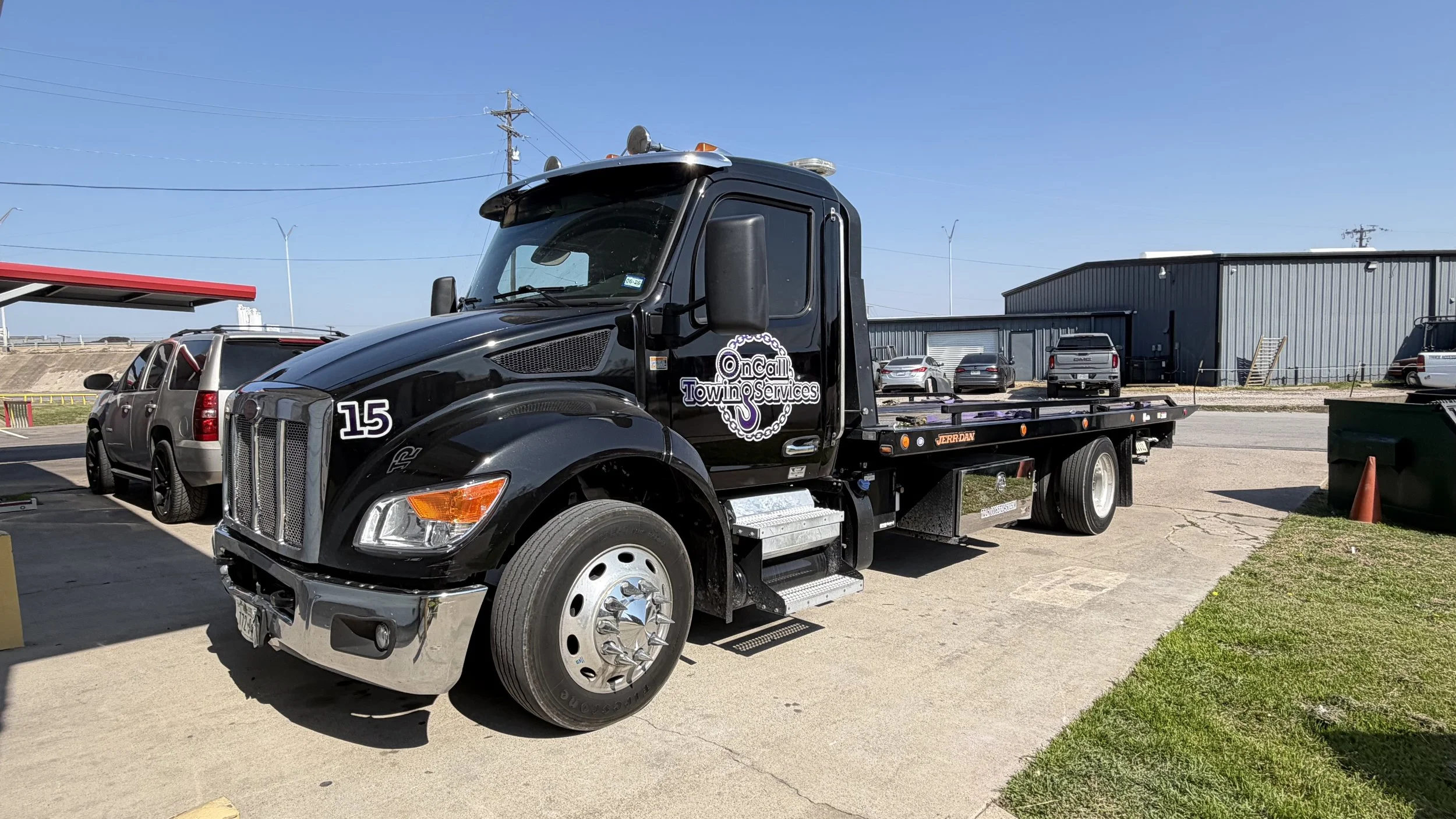 A black tow truck parked in a lot next to other vehicles, with a large warehouse building in the background under a clear blue sky.
