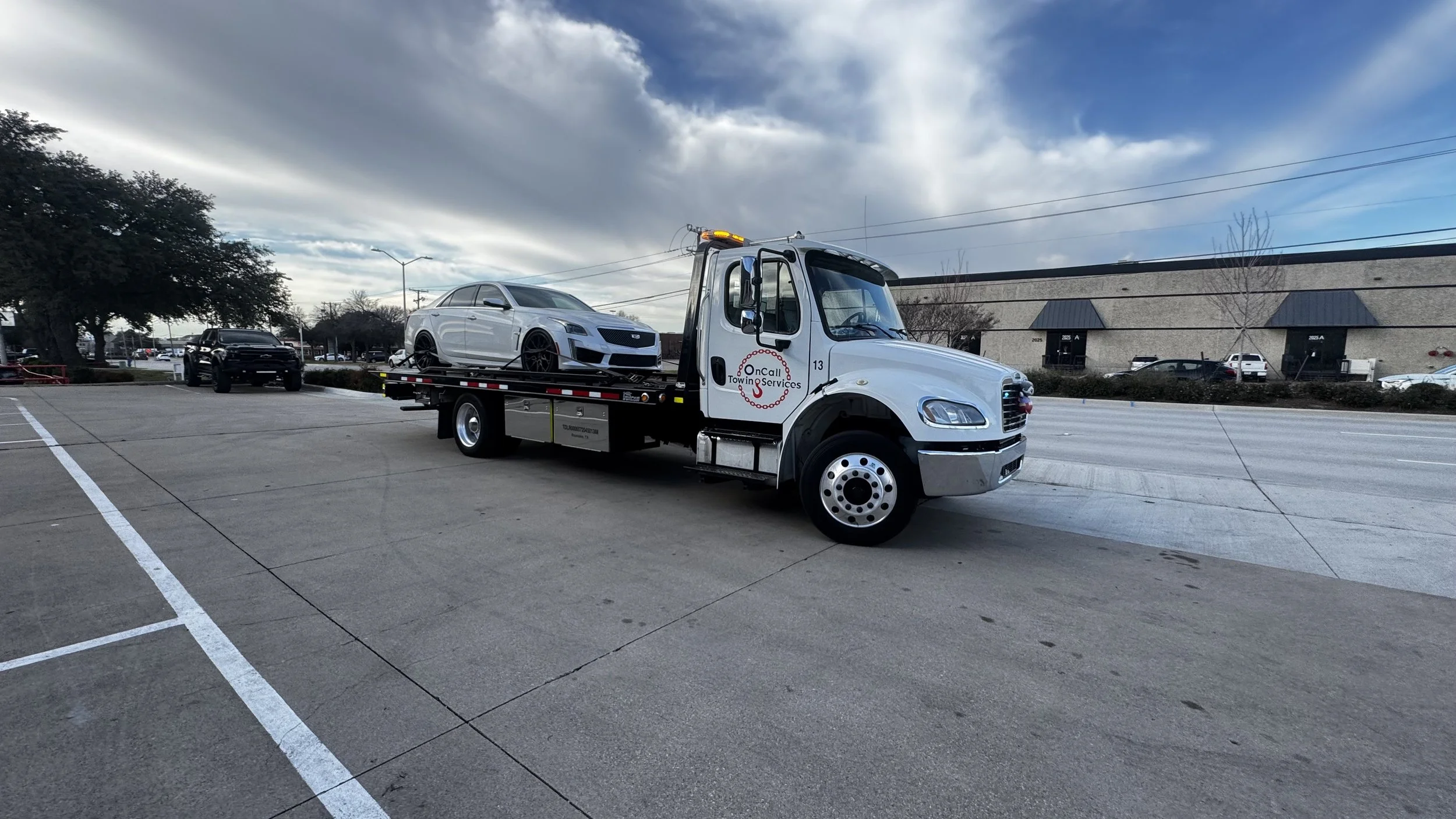 A tow truck with a white luxury car on its flatbed in a parking lot, with trees and buildings in the background under a partly cloudy sky.