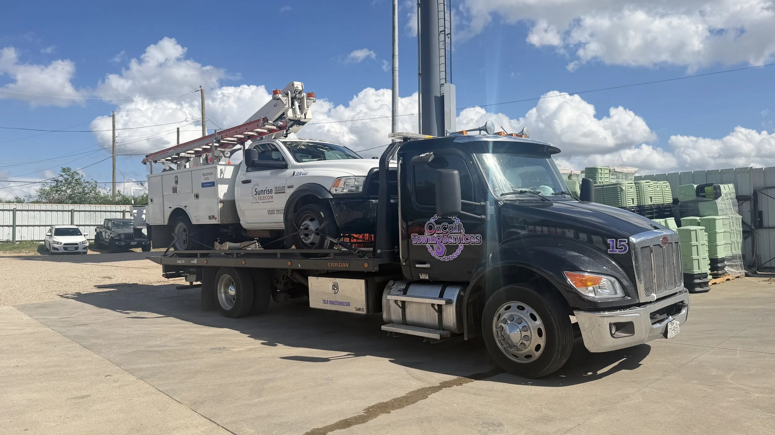 Black tow truck carrying a white utility service vehicle with an extended ladder, parked in an outdoor lot with parked cars and green containers in the background under a partly cloudy sky.