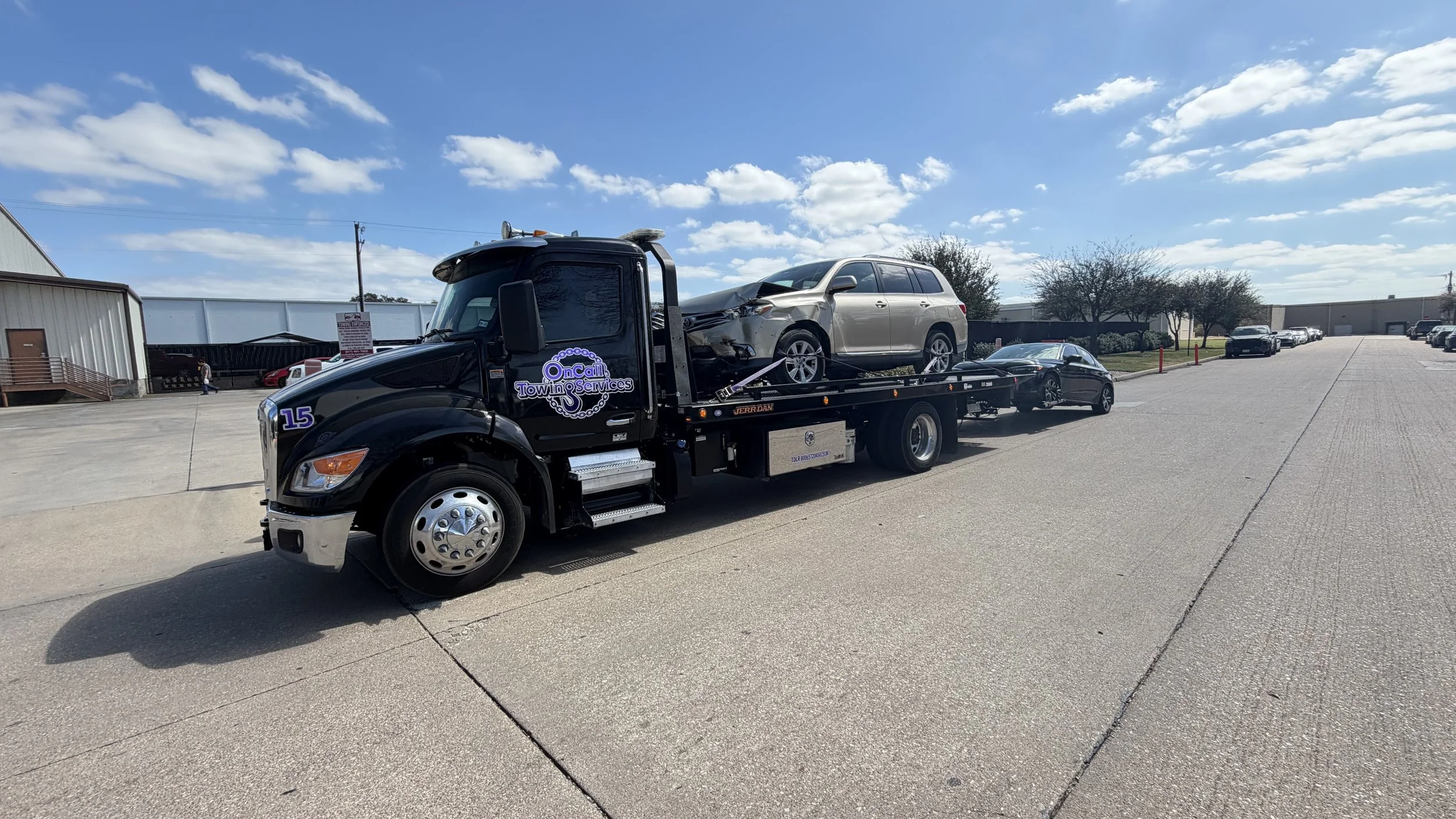 Black tow truck with logo displaying 'OnCall Towing & Services' towing a silver SUV in an open lot, with several other vehicles parked nearby and a partly cloudy sky.