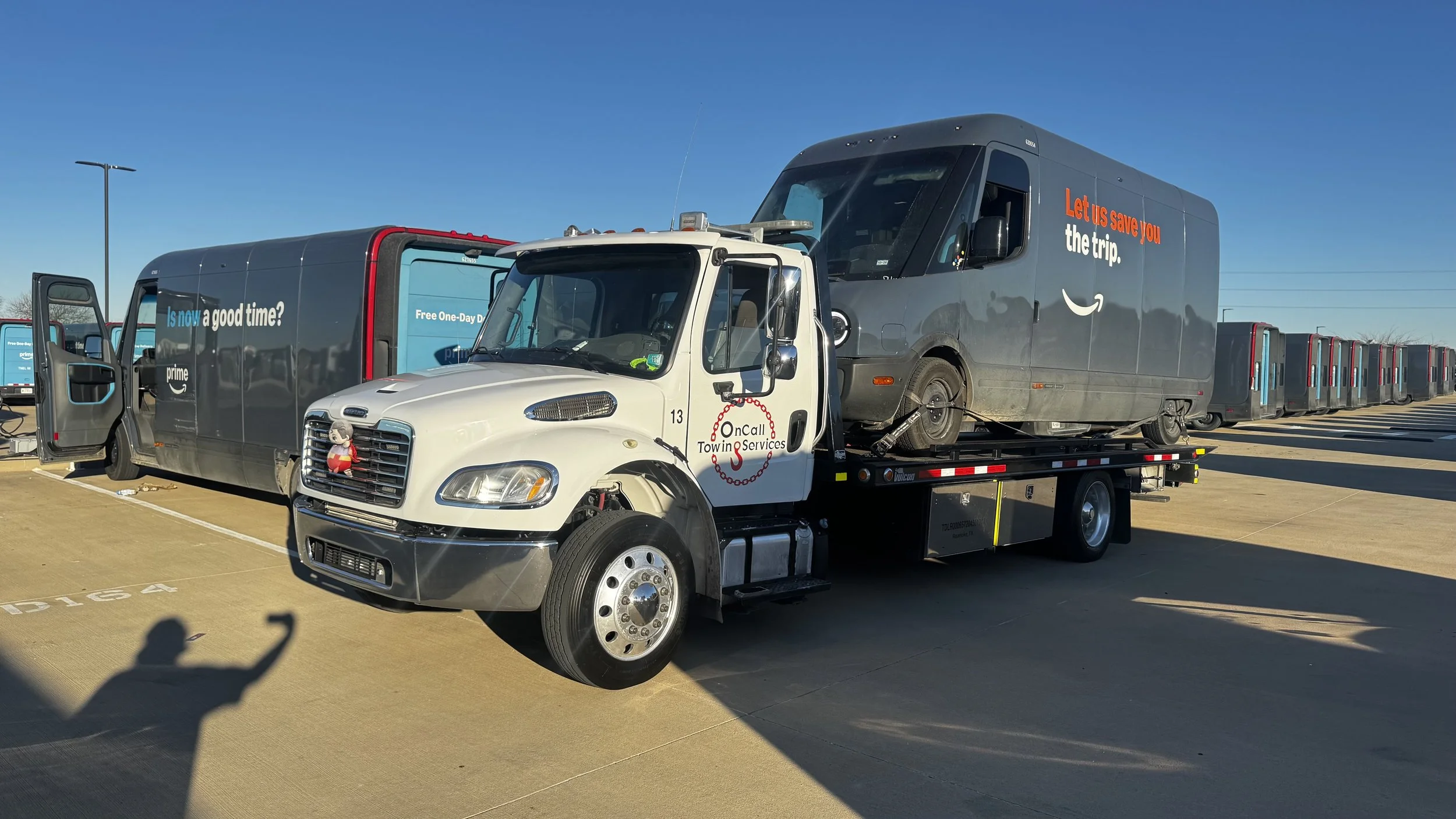 A flatbed tow truck transporting a dark gray Amazon delivery truck in a parking lot, with row of parked Amazon trucks in the background and clear blue sky overhead.