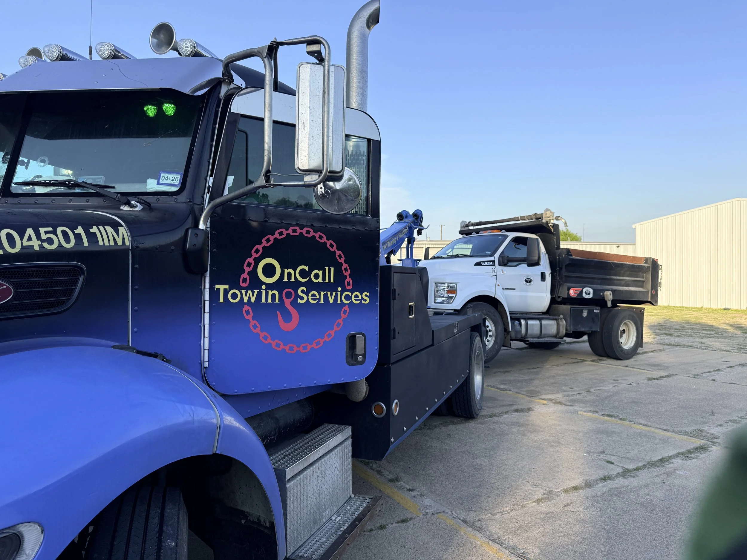 A tow truck with a bumper sticker that reads 'OnCall Towing Services' is towing a white pickup truck. The tow truck is blue with a red chain graphic and a hook logo, parked on a concrete lot with a warehouse in the background under a clear blue sky.