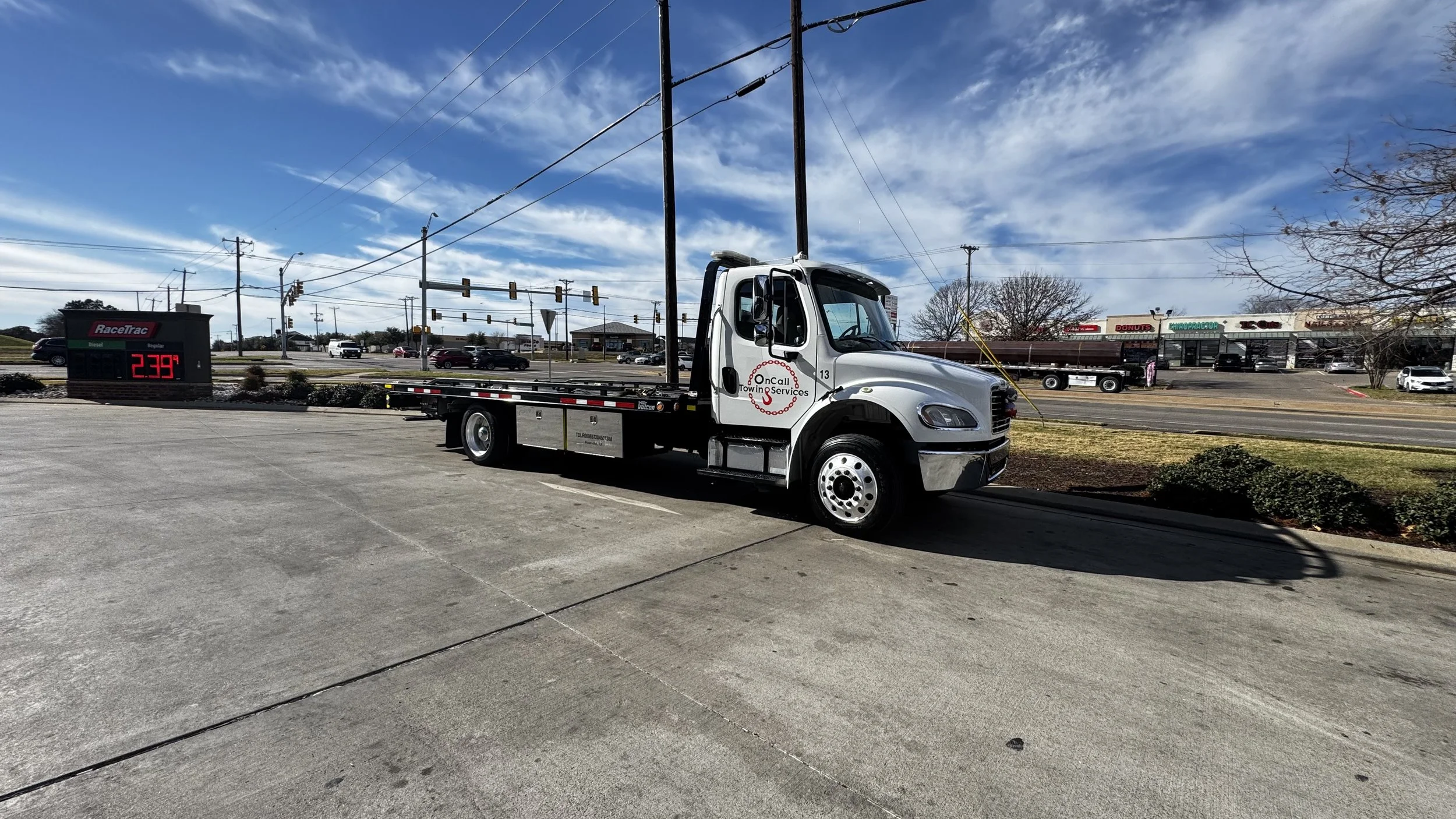 A flatbed tow truck parked on pavement near a sign showing the temperature as 23.9°F, with a shopping center in the background and a blue sky with clouds overhead.