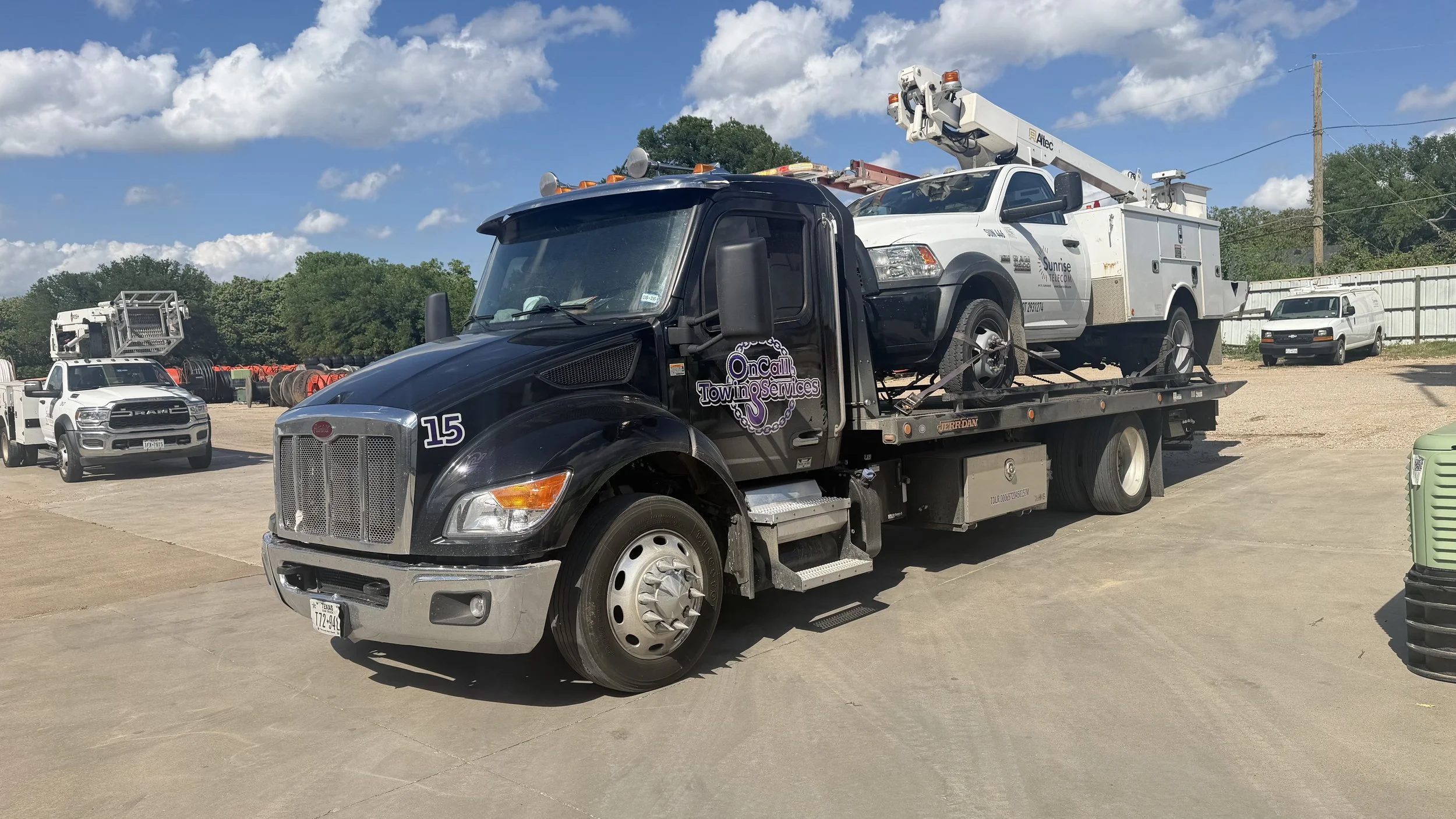 A tow truck carrying a utility vehicle with a bucket lift in a lot with other trucks and vehicles, under a partly cloudy sky.