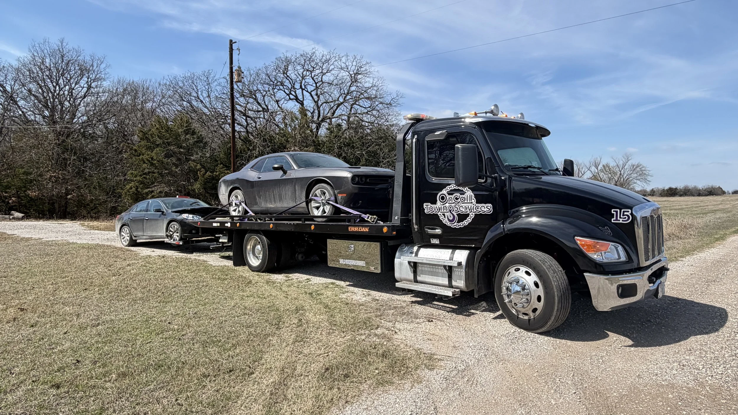 A black tow truck carrying two black cars, with a third car parked nearby on a gravel path. The scene is outdoors with trees and a blue sky in the background.