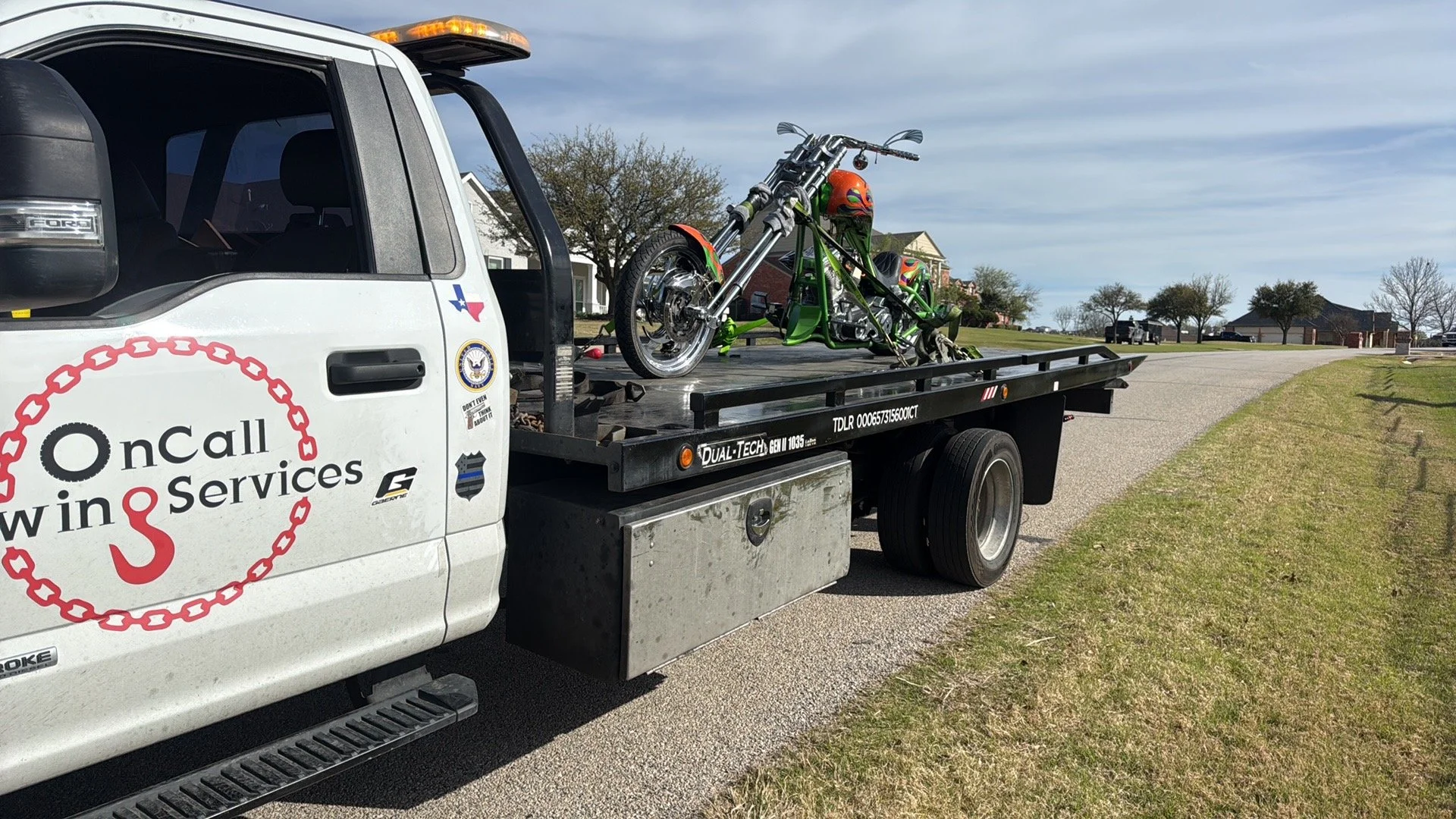A flatbed tow truck parked on the side of a suburban street, carrying a green and chrome motorcycle with orange and black helmet on it.
