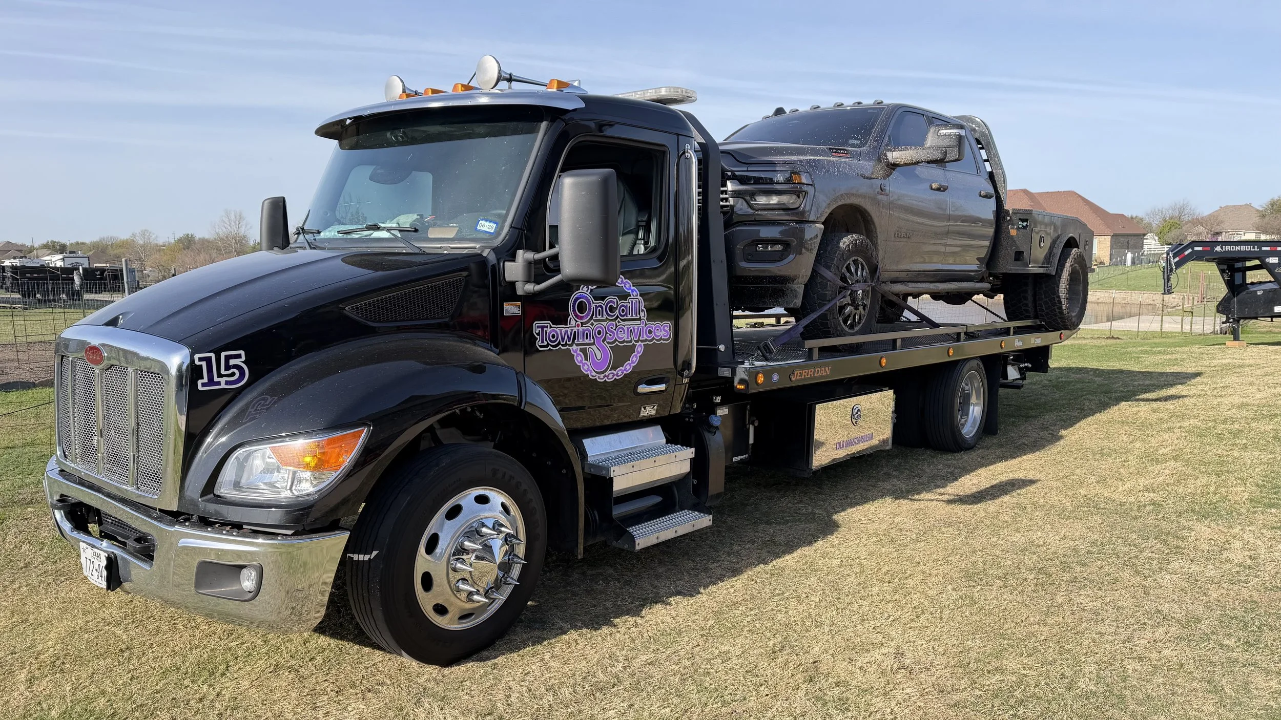 Black tow truck with purple lettering on door, towing a black pickup truck on a grassy field with houses and trees in the background.