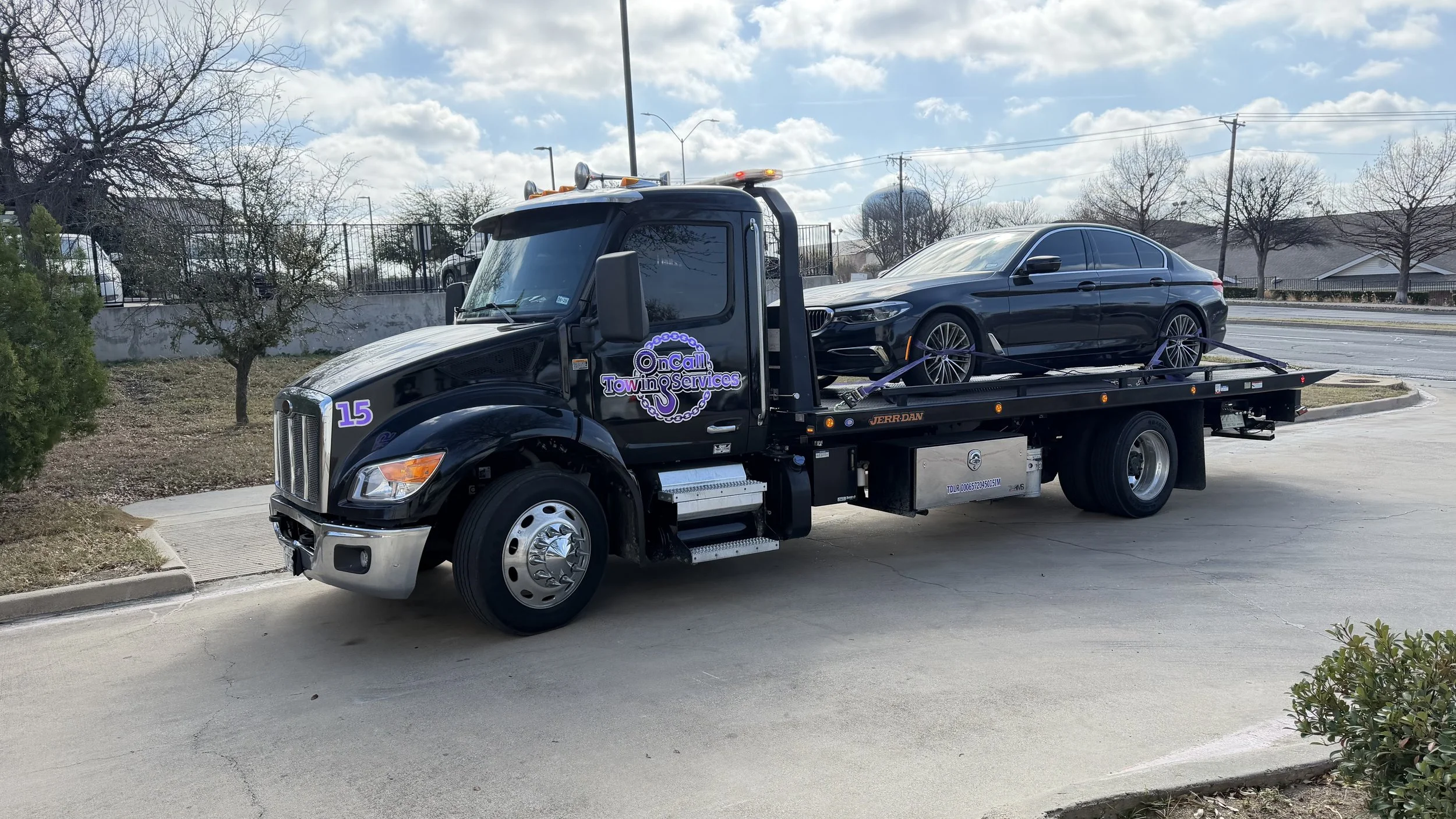 A tow truck transporting a black sedan, with the tow truck displaying a logo that reads "On Call Tow & Services" on the door.
