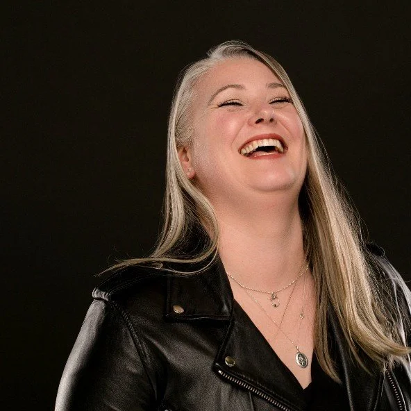 Woman with long blonde hair laughing, wearing a black leather jacket and layered necklaces, against a dark background.