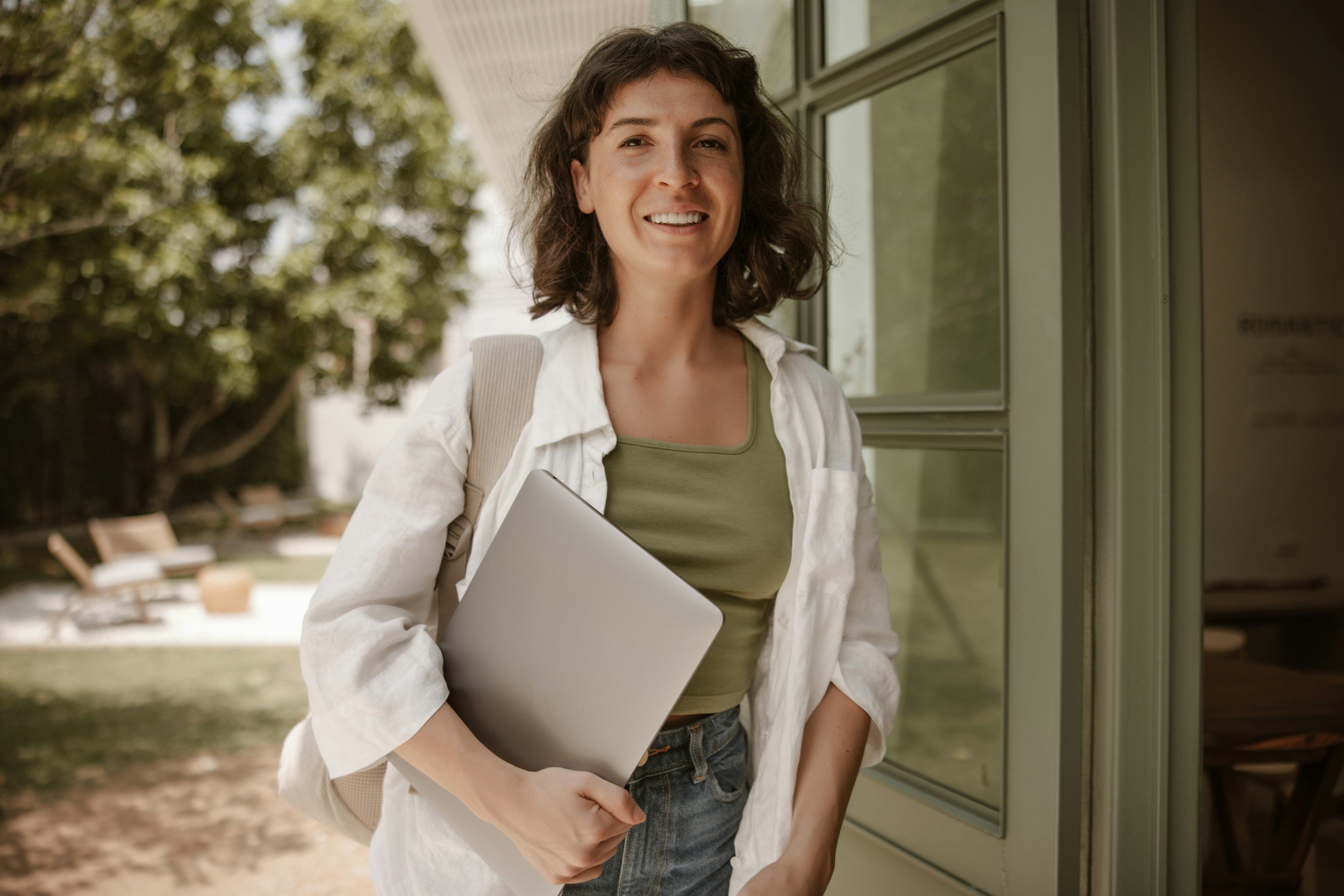 A young woman with short brown hair smiling, holding a silver laptop, dressed in a white shirt and green top, standing outside near a glass door with trees and outdoor furniture in the background.